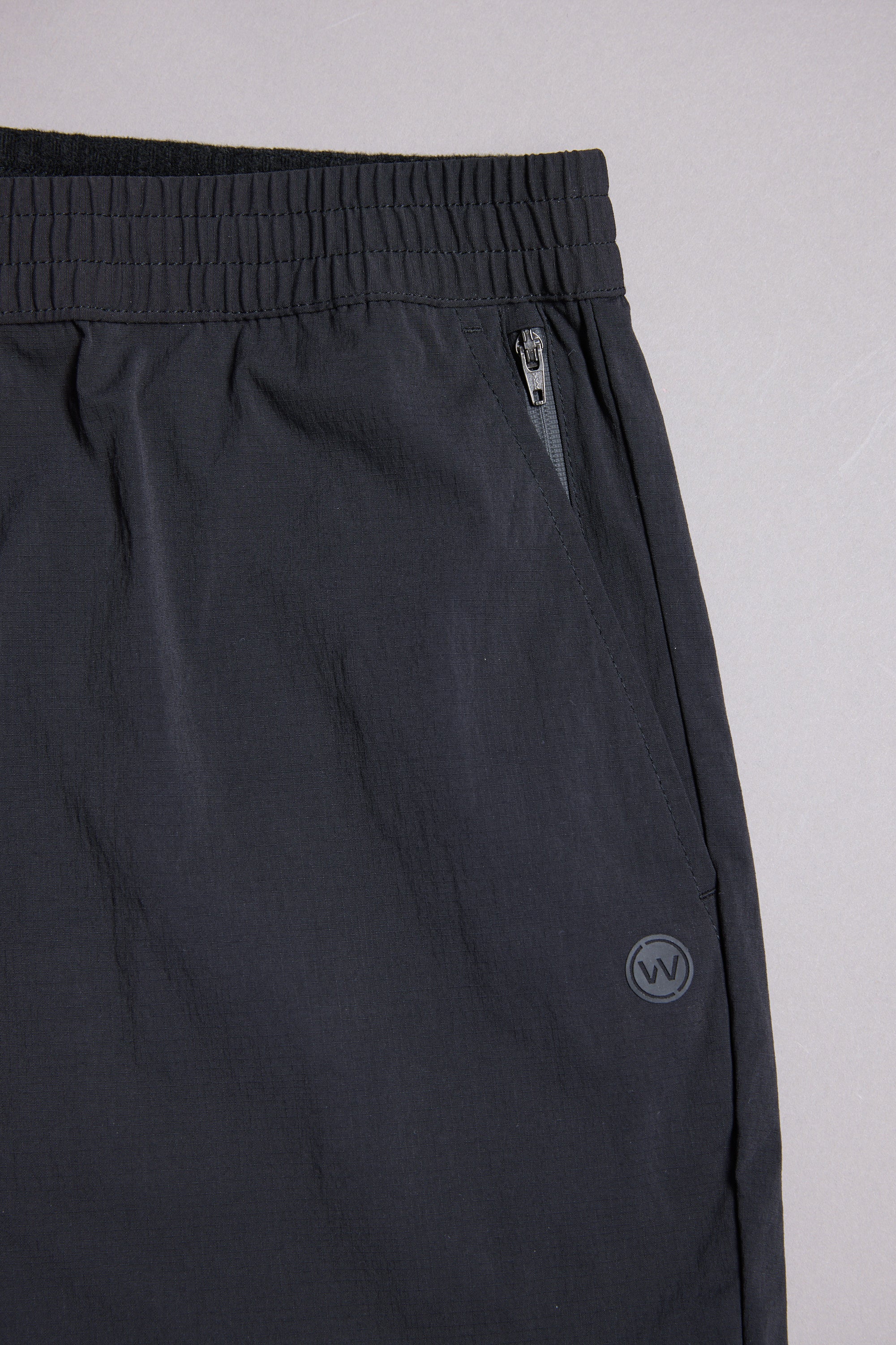 Close-up of WearEver Transit Pant, featuring an elastic waistband, zippered pocket, and small circular logo on the right side near the pocket. Shown against a neutral gray background.