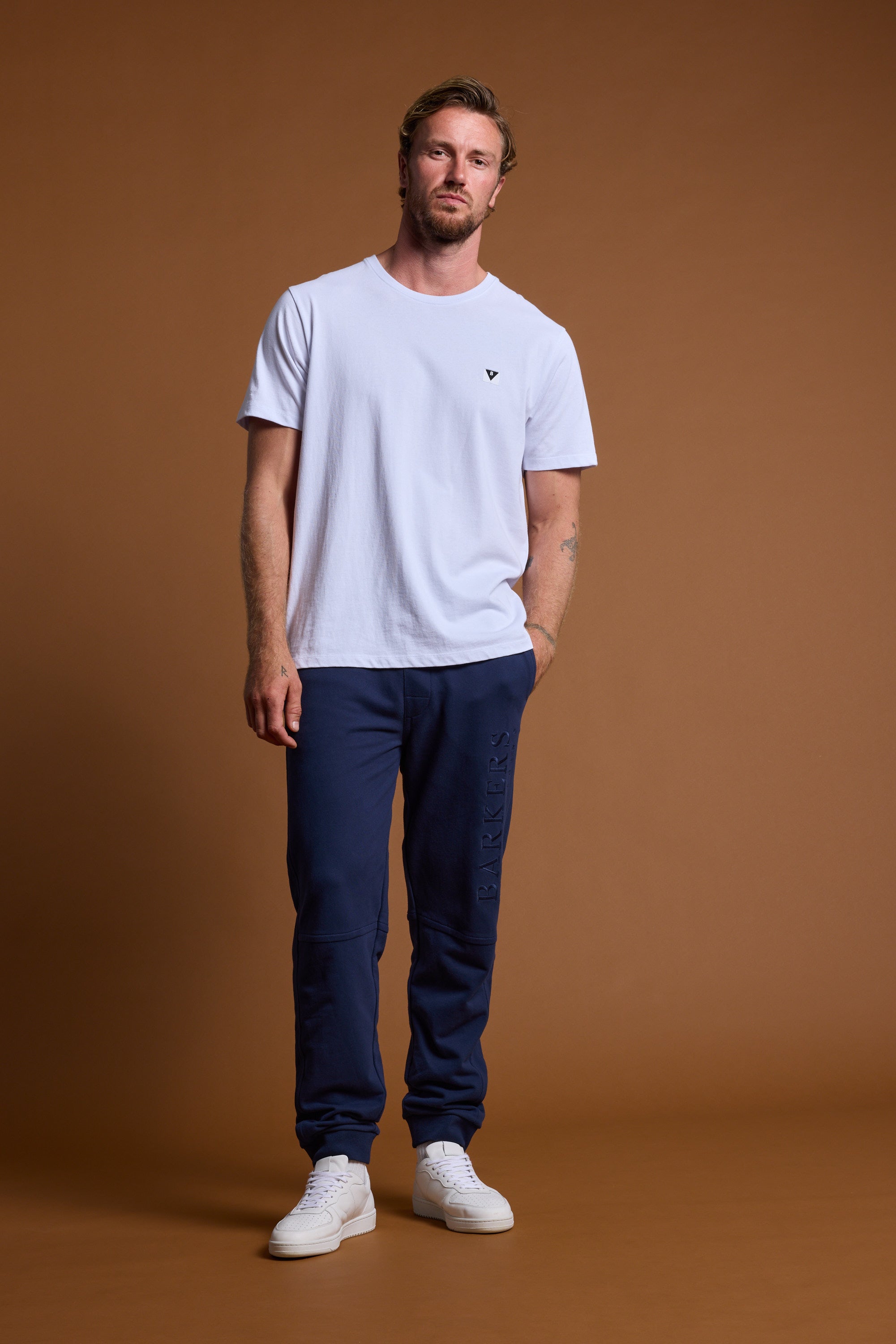 A man stands against a brown background, wearing a plain white T-shirt, Barkers Heritage Track Pant by Barkers in navy blue French terry, and white sneakers. He poses casually with one hand in his pocket.