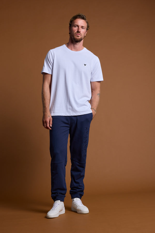 A man stands against a brown background, wearing a plain white T-shirt, Barkers Heritage Track Pant by Barkers in navy blue French terry, and white sneakers. He poses casually with one hand in his pocket.