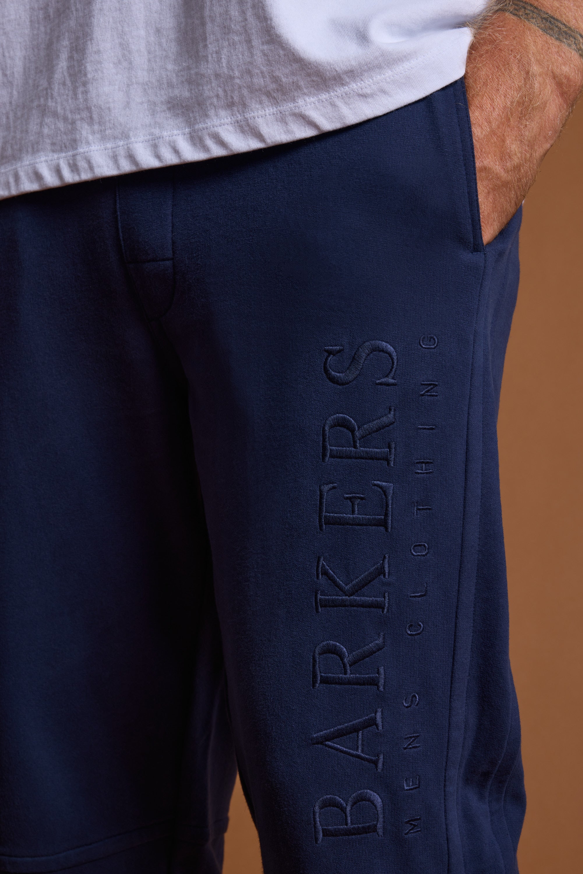 Close-up of a person wearing Barkers Heritage Track Pant in navy blue with Barkers branding on the leg and a white shirt visible. Their hand is in the pocket, set against a brown background.