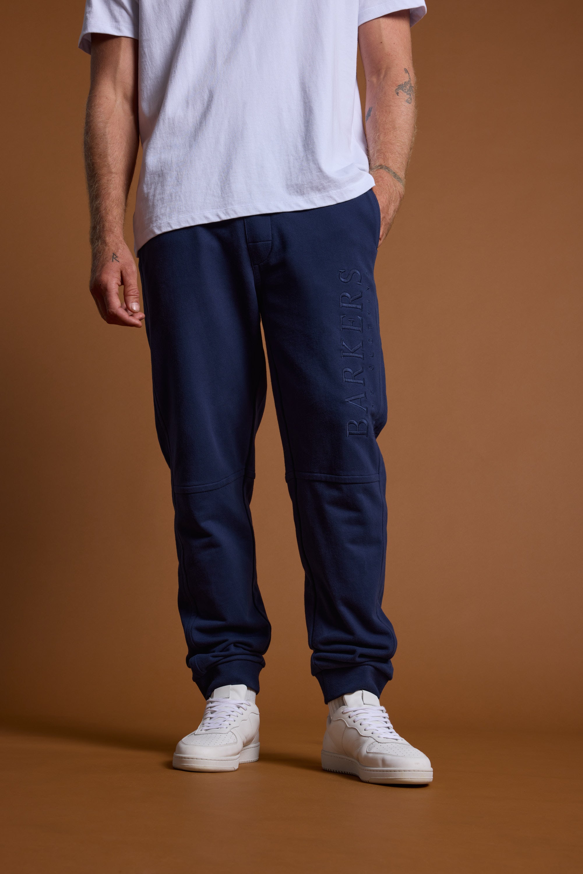 A person stands against a brown background, wearing white sneakers and Barkers Heritage Track Pant by Barkers in navy blue organic cotton with "BARKERS" embossed on the leg. The upper body is mostly out of frame.