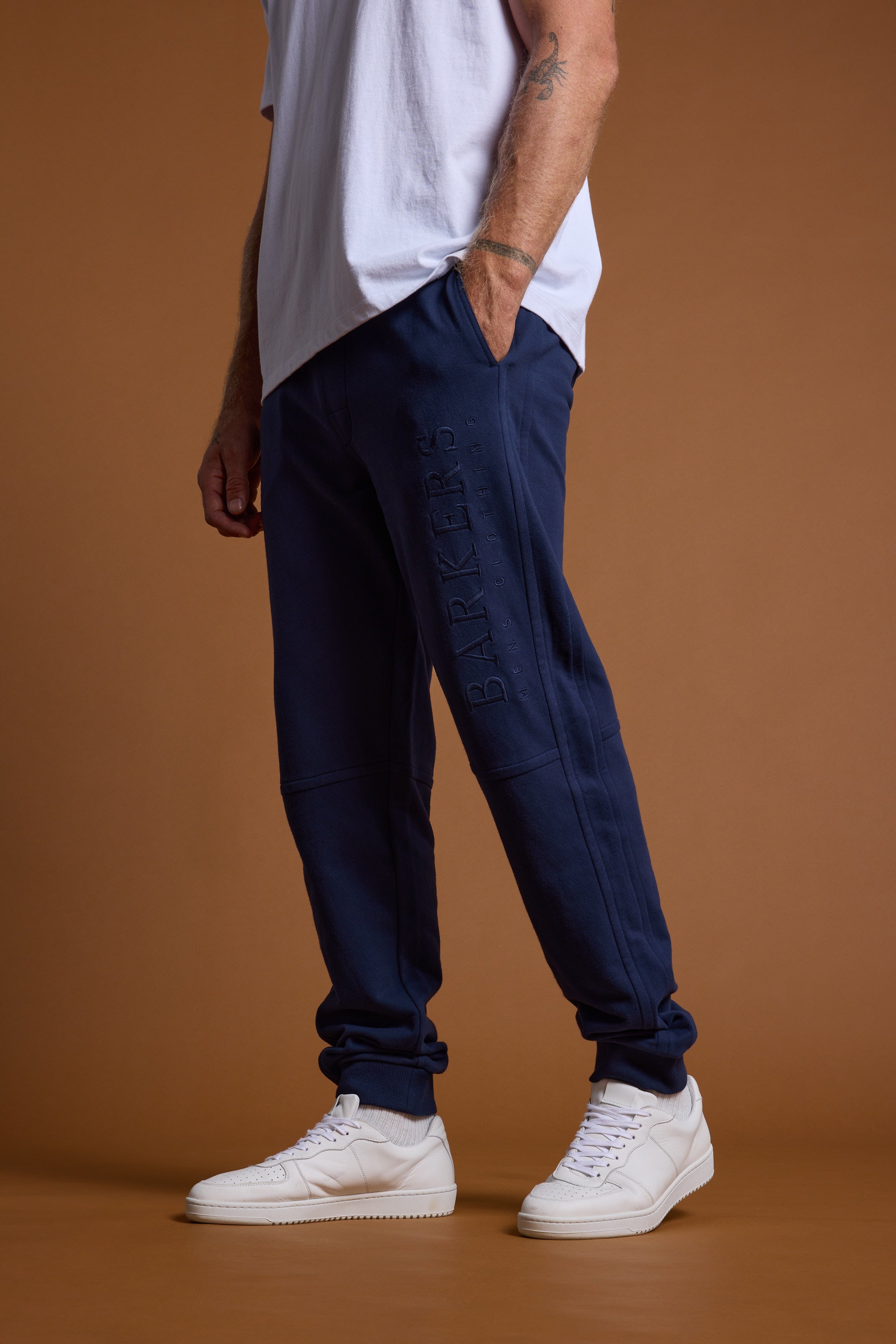 A person wearing a white T-shirt, Barkers Heritage Track Pant in navy blue with BARKERS text on the side, and white sneakers stands against a plain brown background.
