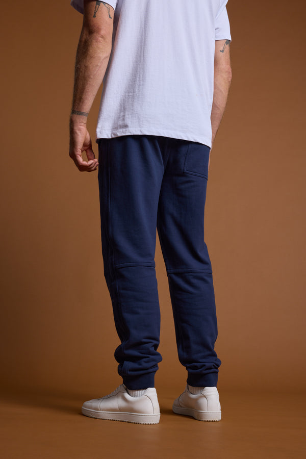 Only the lower half of a person is visible from the back against a brown background, wearing Barkers Heritage Track Pant in navy blue by Barkers, styled with a white T-shirt and white sneakers.