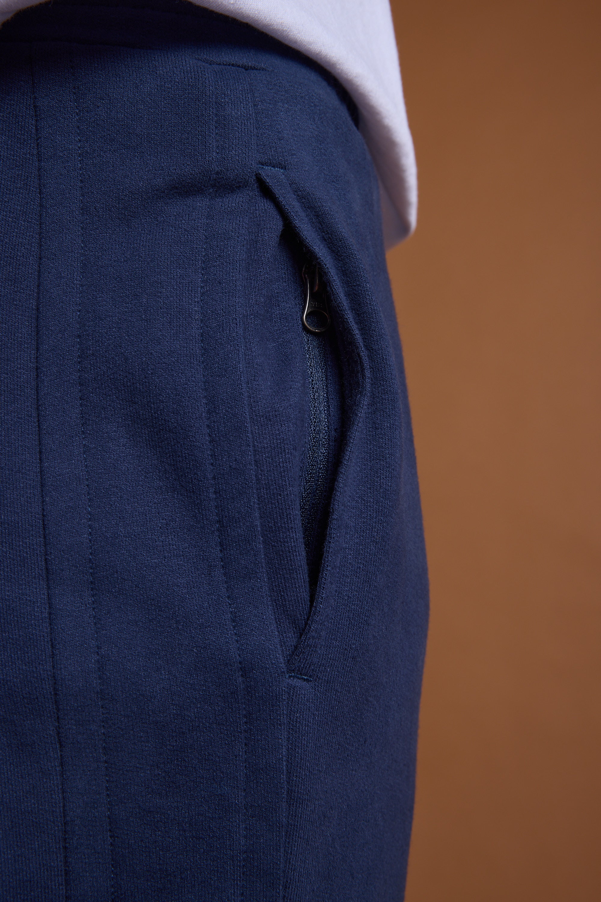 Close-up of a person in navy blue Barkers Heritage Track Pant by Barkers, featuring a zippered pocket and a white shirt partially tucked at the waist, set against a solid brown background.