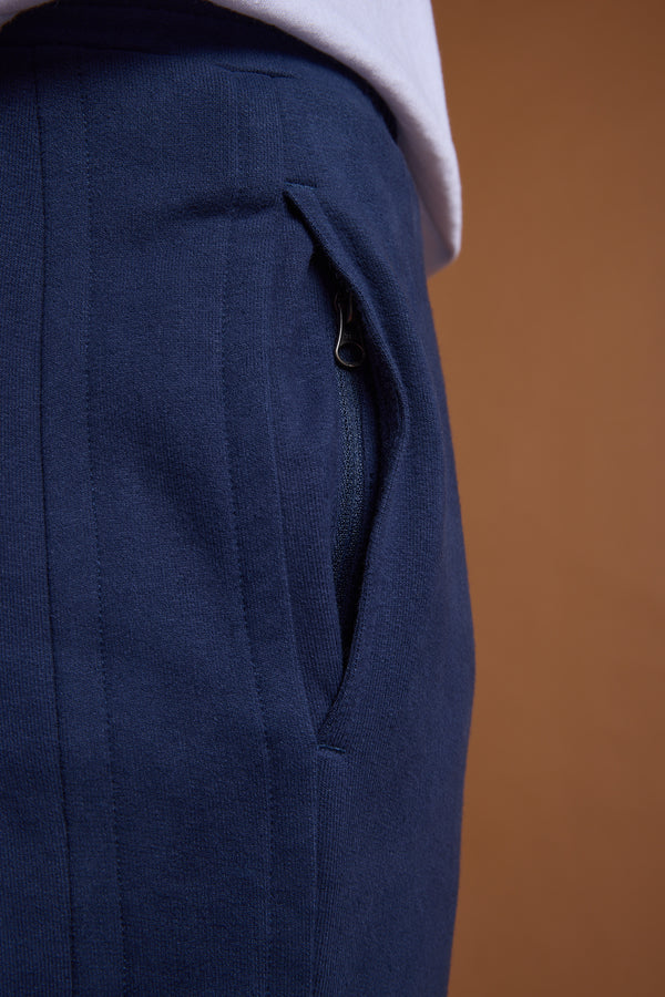 Close-up of a person in navy blue Barkers Heritage Track Pant by Barkers, featuring a zippered pocket and a white shirt partially tucked at the waist, set against a solid brown background.