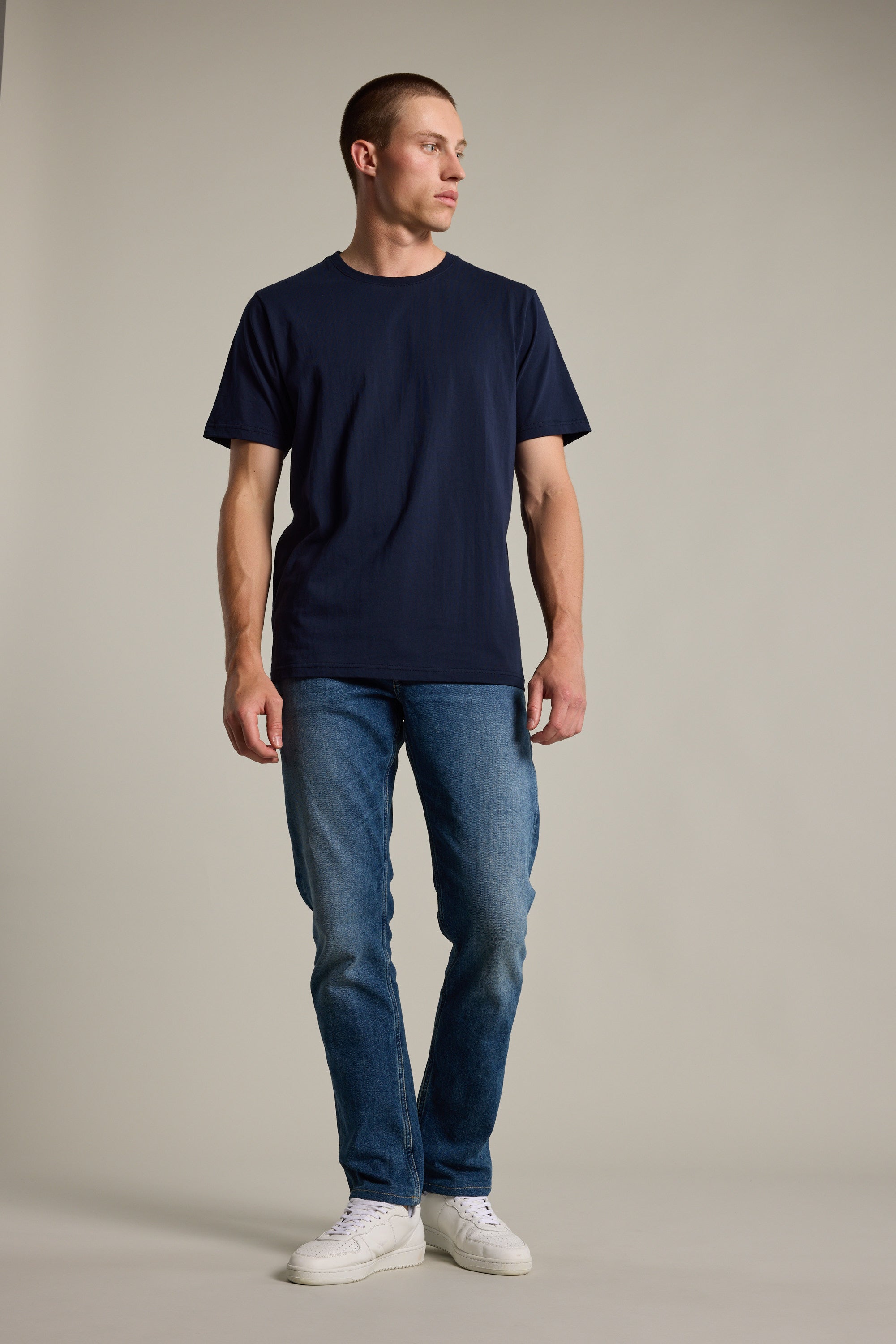 A man stands against a plain background, wearing Barkers' navy blue Organic Crew Tee made from 100% organic cotton, paired with blue jeans and white sneakers, looking slightly left with his hands relaxed at his sides.