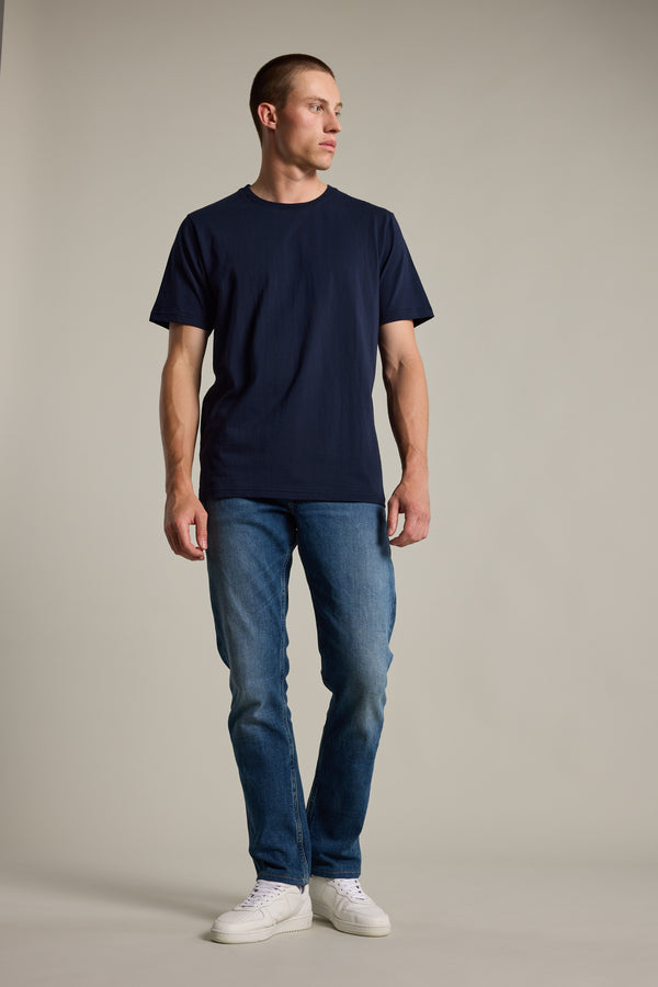 A man stands against a plain background, wearing Barkers' navy blue Organic Crew Tee made from 100% organic cotton, paired with blue jeans and white sneakers, looking slightly left with his hands relaxed at his sides.