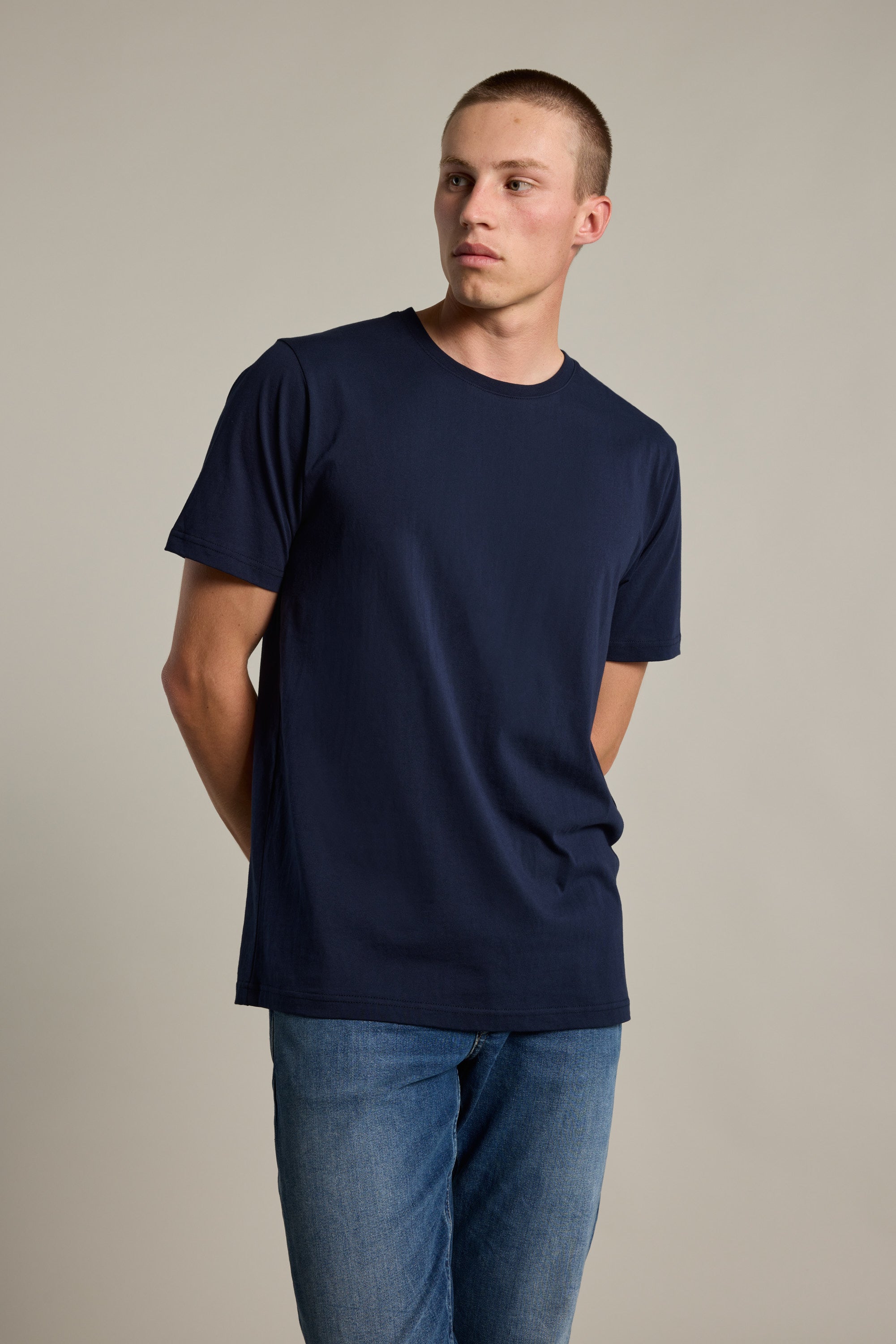 A young man wears the Barkers Organic Crew Tee, made from 100% organic cotton in navy blue, paired with blue jeans. He stands against a neutral background, looking slightly left with his hands behind his back.