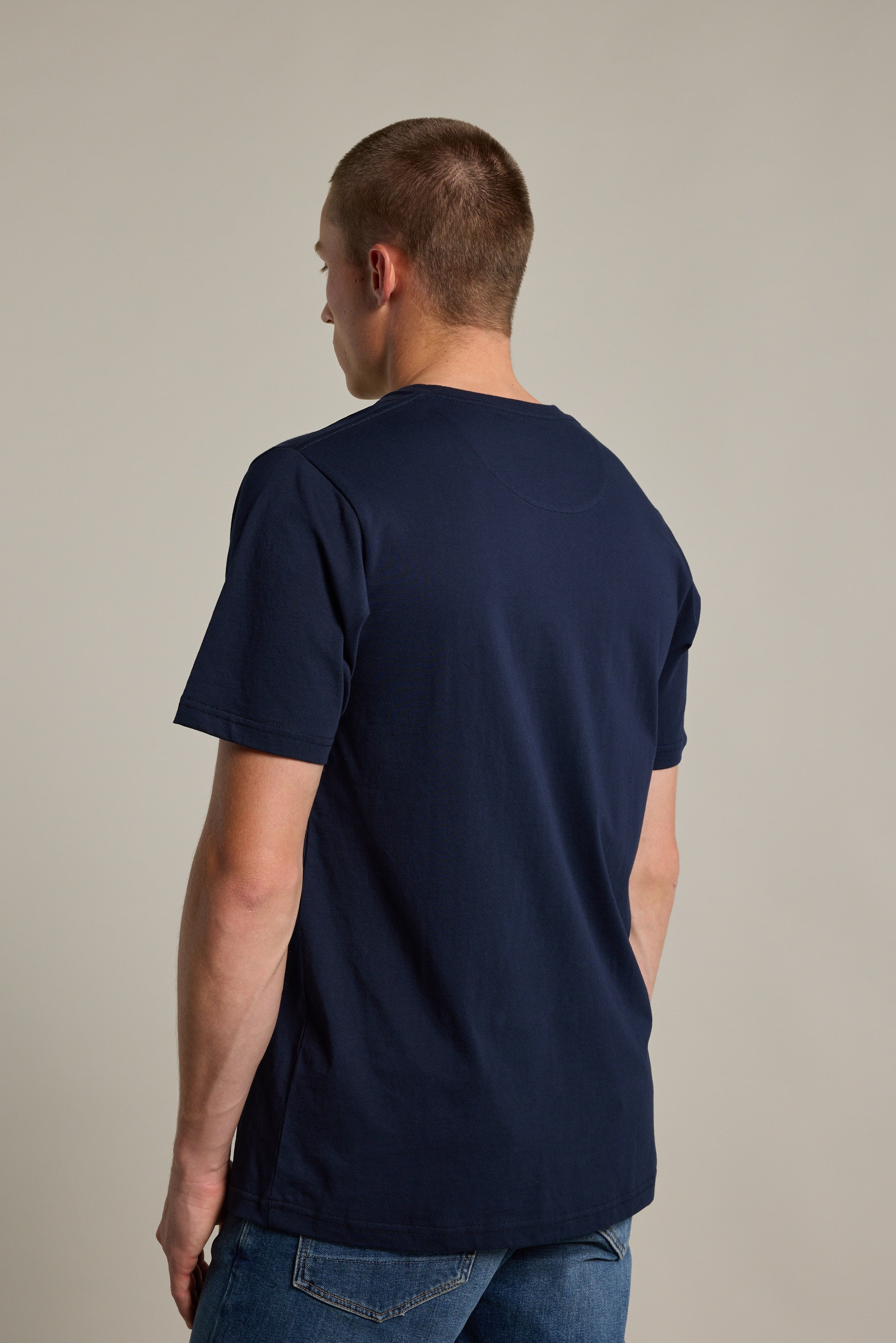 A man with short hair, wearing Barkers' Organic Crew Tee in navy blue and blue jeans, stands facing away from the camera against a plain light background.