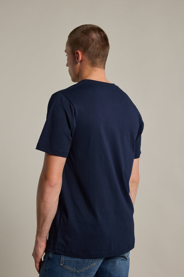 A man with short hair, wearing Barkers' Organic Crew Tee in navy blue and blue jeans, stands facing away from the camera against a plain light background.