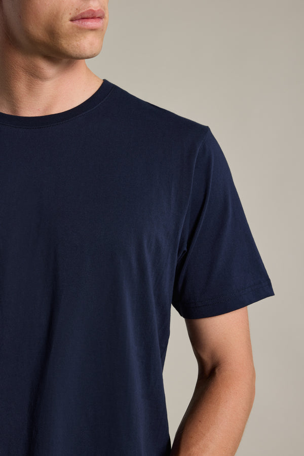 A man models the Barkers Organic Crew Tee in plain navy blue, crafted from 100% organic cotton; only his lower face, upper torso, and right arm are visible against a neutral background.