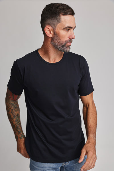 A man with short dark hair and a beard, wearing the Barkers Linton Supima Tee in black and light blue jeans, stands against a neutral background looking left. A colorful tattoo is visible on his arm—an ideal wardrobe staple.