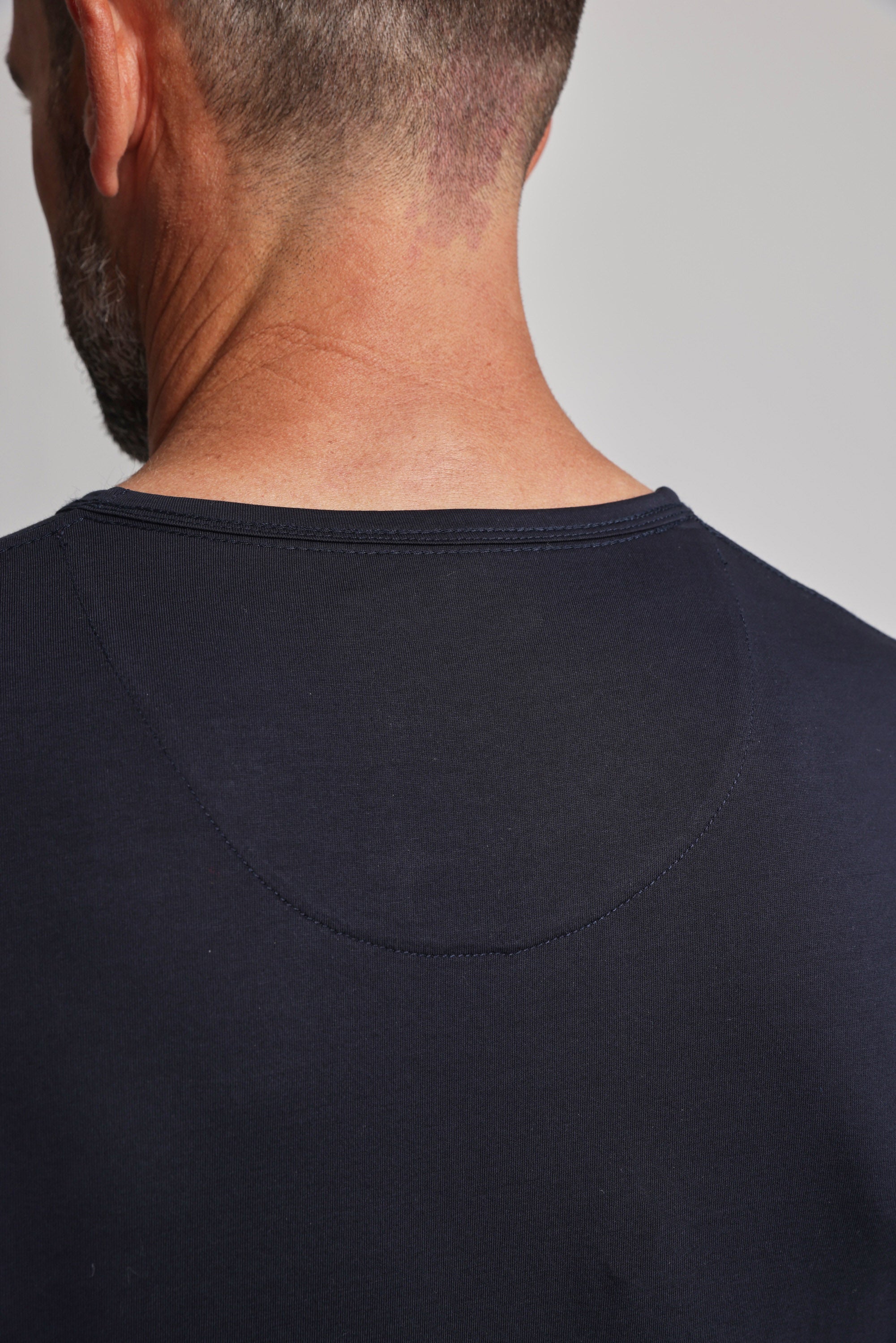 The image shows the back view of a man with short hair wearing the Barkers Linton Supima Tee, a dark navy blue crew neck made from mercerised Supima cotton, highlighting its neckline and stitching details.