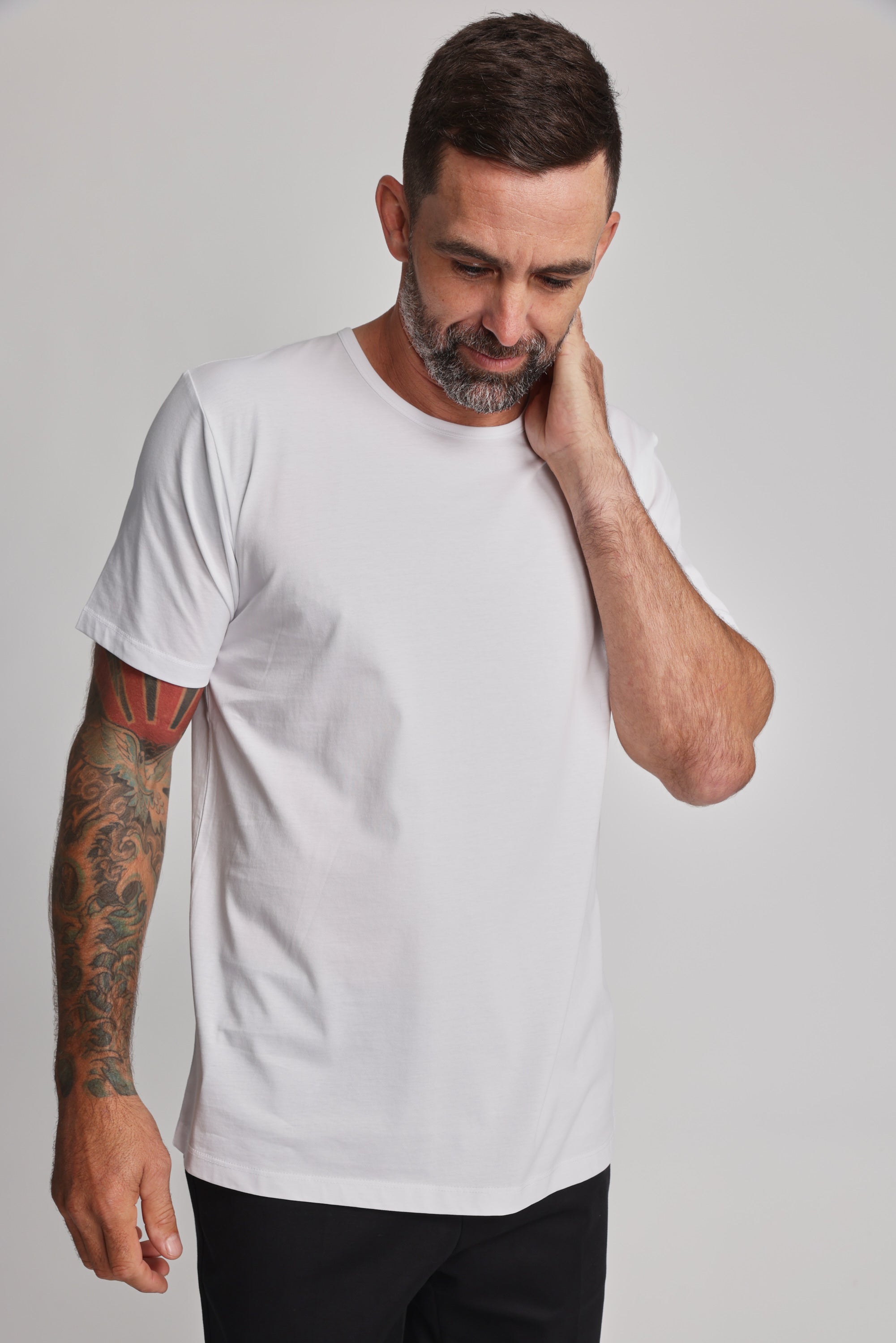 A man with short dark hair and a beard wears the Barkers Linton Supima Tee and black pants. He looks down, touching his neck, showing a colorful tattoo sleeve on his left arm; the background is plain and light-colored.