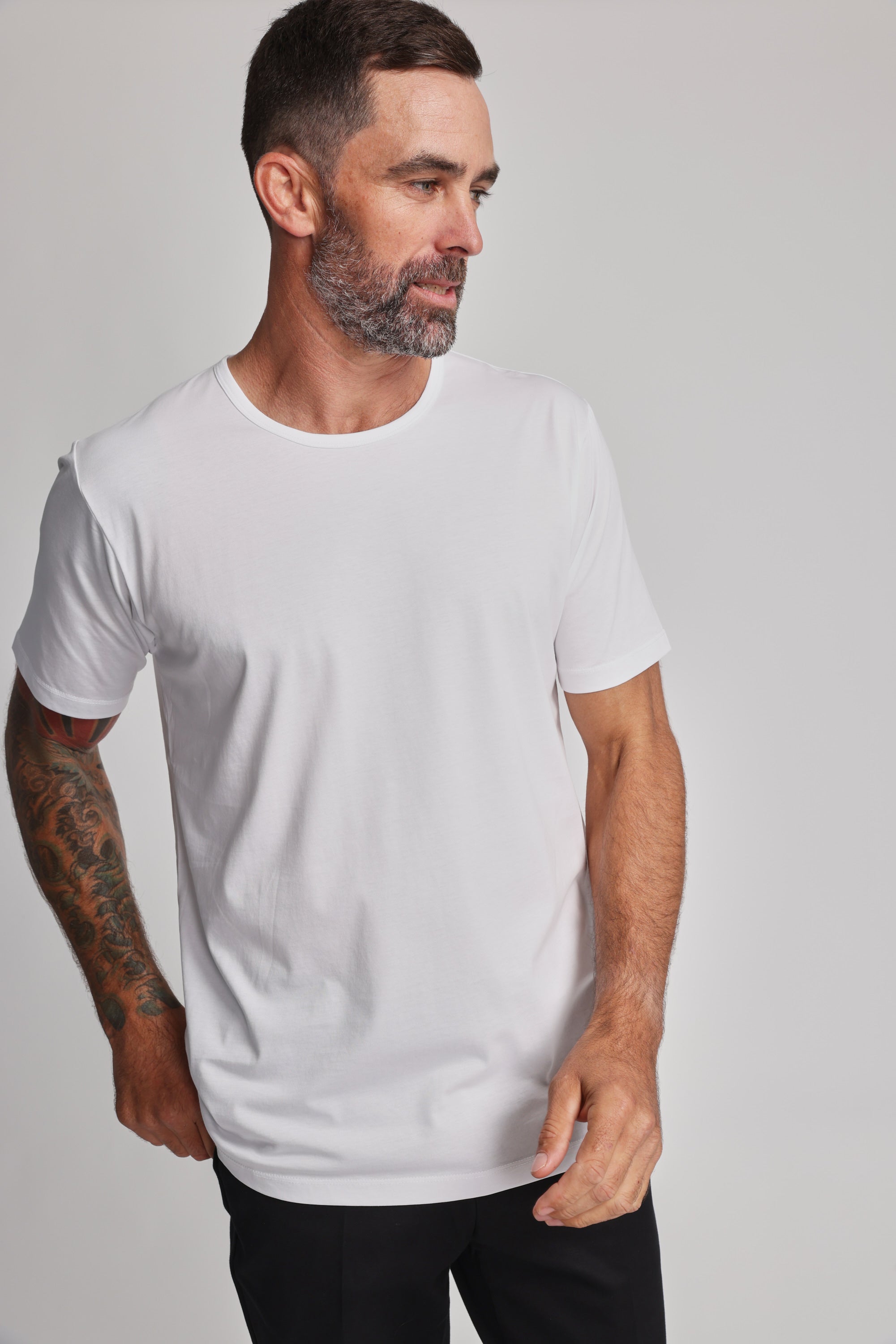 A man with short dark hair, a beard, and a tattoo sleeve on his left arm wears the Barkers Linton Supima Tee in white with black pants. He faces left with a neutral expression against a light background.