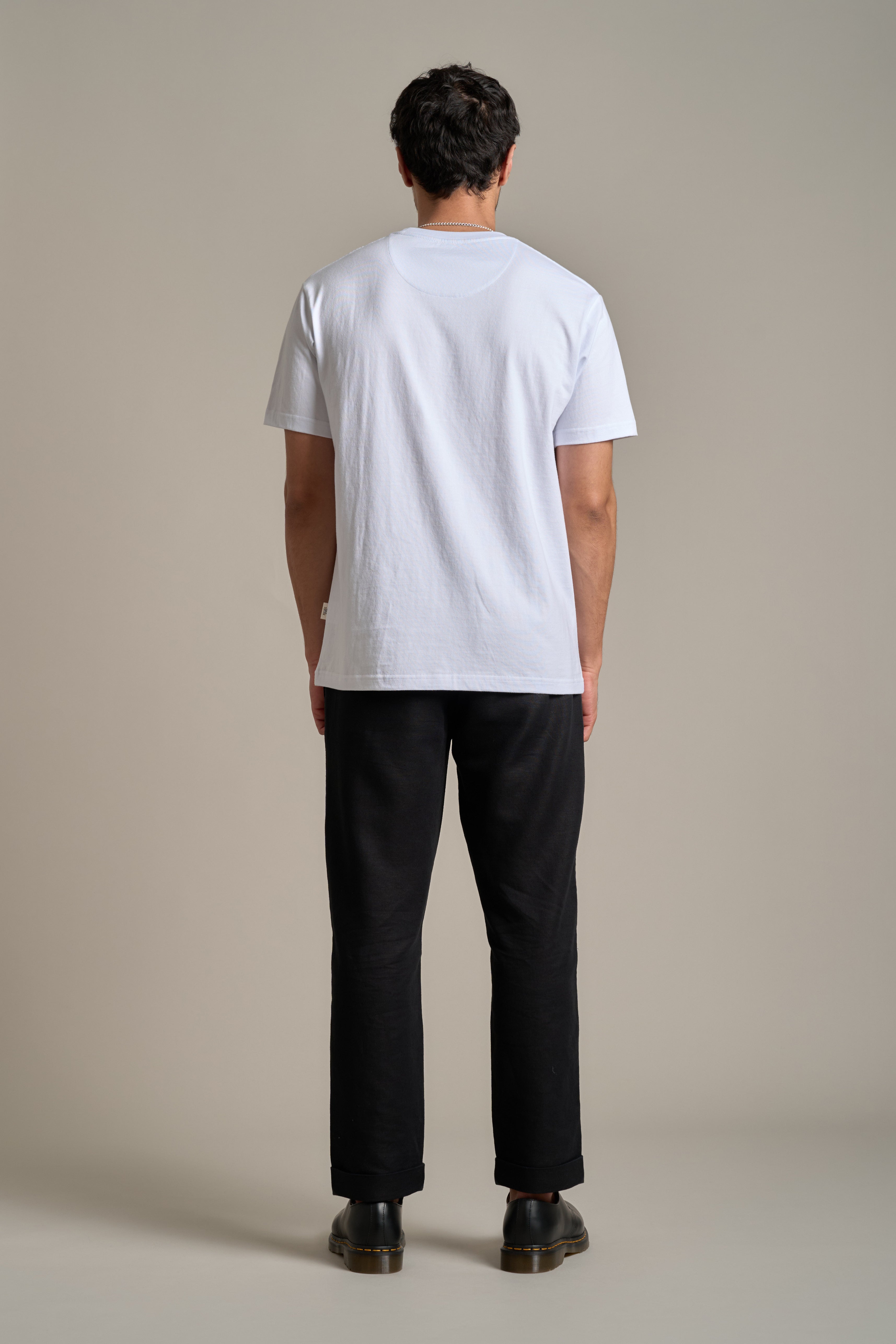 A person faces away from the camera, wearing the Barkers Boardriders Logo Tee in white, paired with black pants and shoes against a neutral background—an everyday essential look.