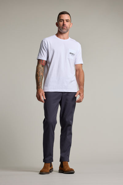 A man with short hair and right arm tattoos stands against a plain background, wearing a Barkers Basecamp Logo Tee by Barkers, dark gray pants, and brown suede boots—a staple for any Kiwi wardrobe.