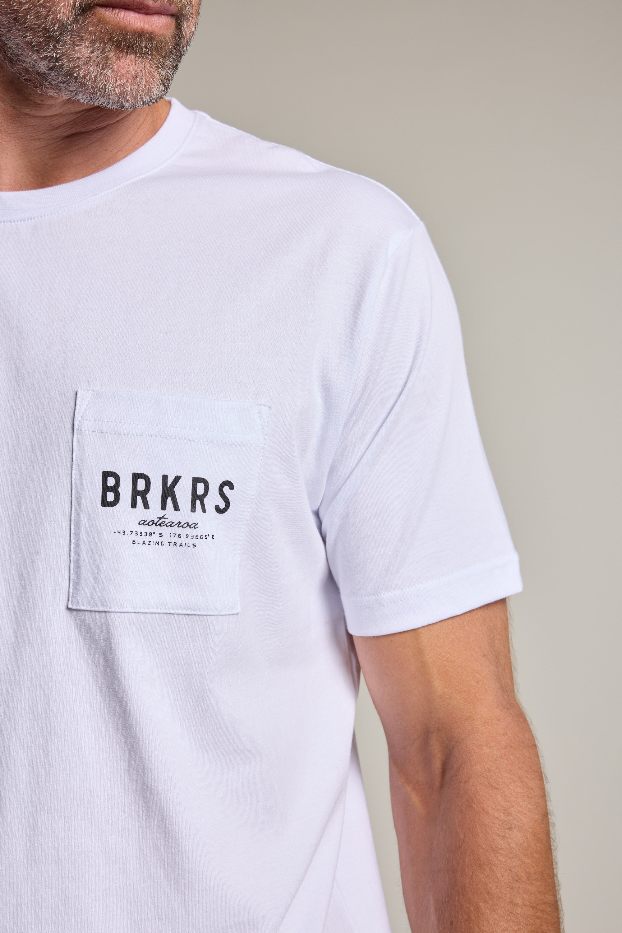 A man wears the Barkers Basecamp Logo Tee by Barkers, a white organic cotton T-shirt with a chest pocket and “BRKRS authentic” print. Only his lower face and upper body are shown—an essential piece for any Kiwi wardrobe.