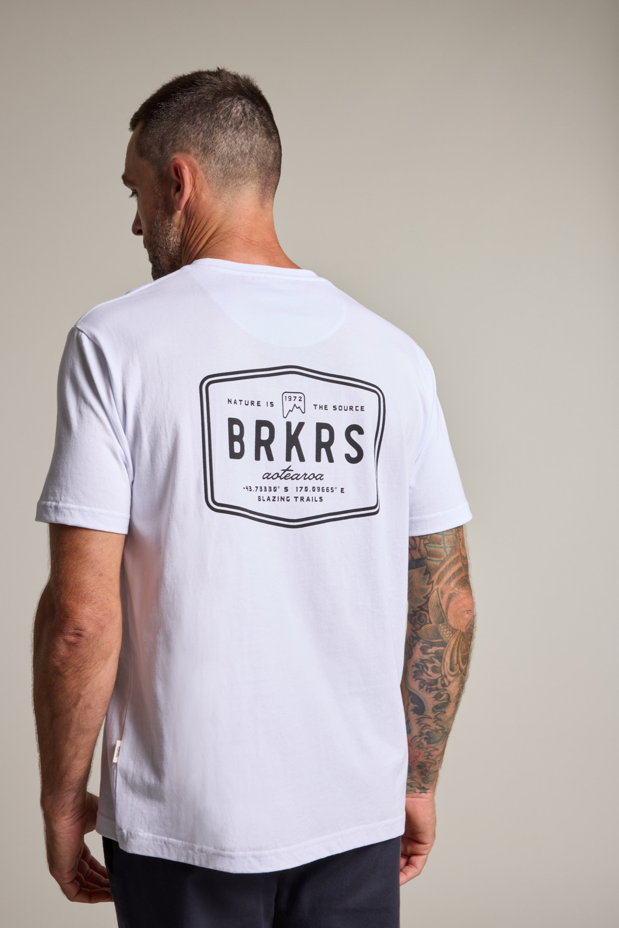 A man with short hair and a tattooed right arm wears the Barkers Basecamp Logo Tee by Barkers, made from organic cotton and featuring a bold black and white logo on the back—an essential for any Kiwi wardrobe.