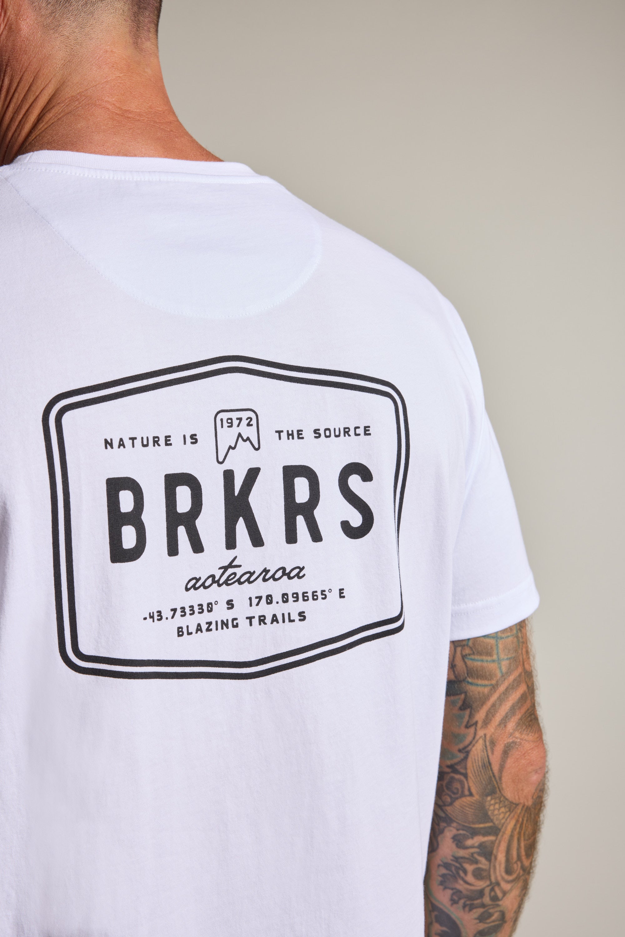 A person wears the Barkers Basecamp Logo Tee by Barkers, made from organic cotton with a bold black BRKRS logo and coordinates on the back. "Nature is the source, Aotearoa, Blazing Trails" gives Kiwi style. A tattooed arm is partially visible.