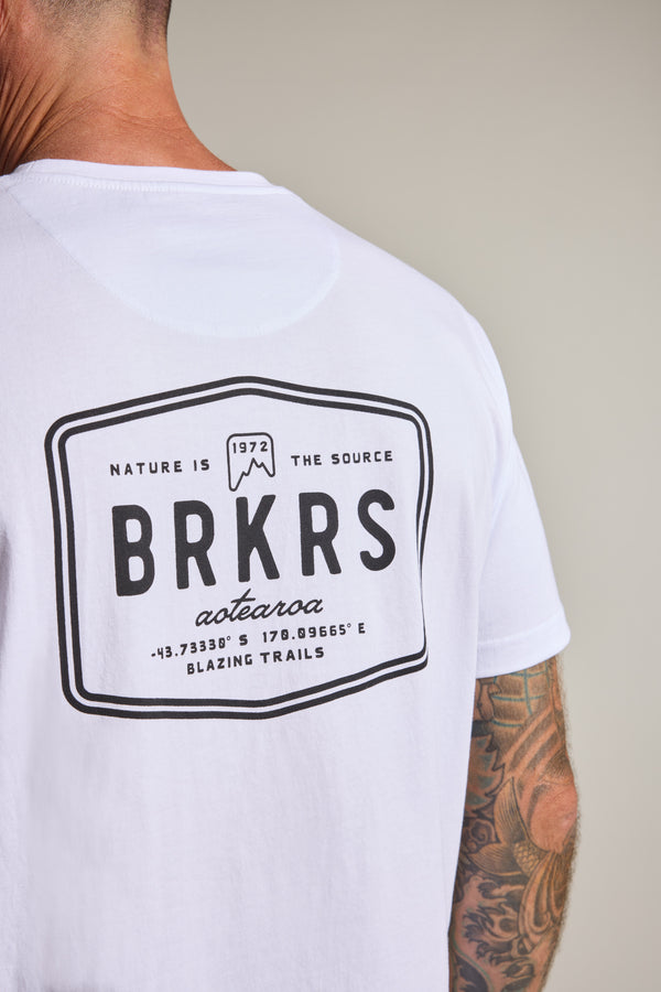 A person wears the Barkers Basecamp Logo Tee by Barkers, made from organic cotton with a bold black BRKRS logo and coordinates on the back. "Nature is the source, Aotearoa, Blazing Trails" gives Kiwi style. A tattooed arm is partially visible.