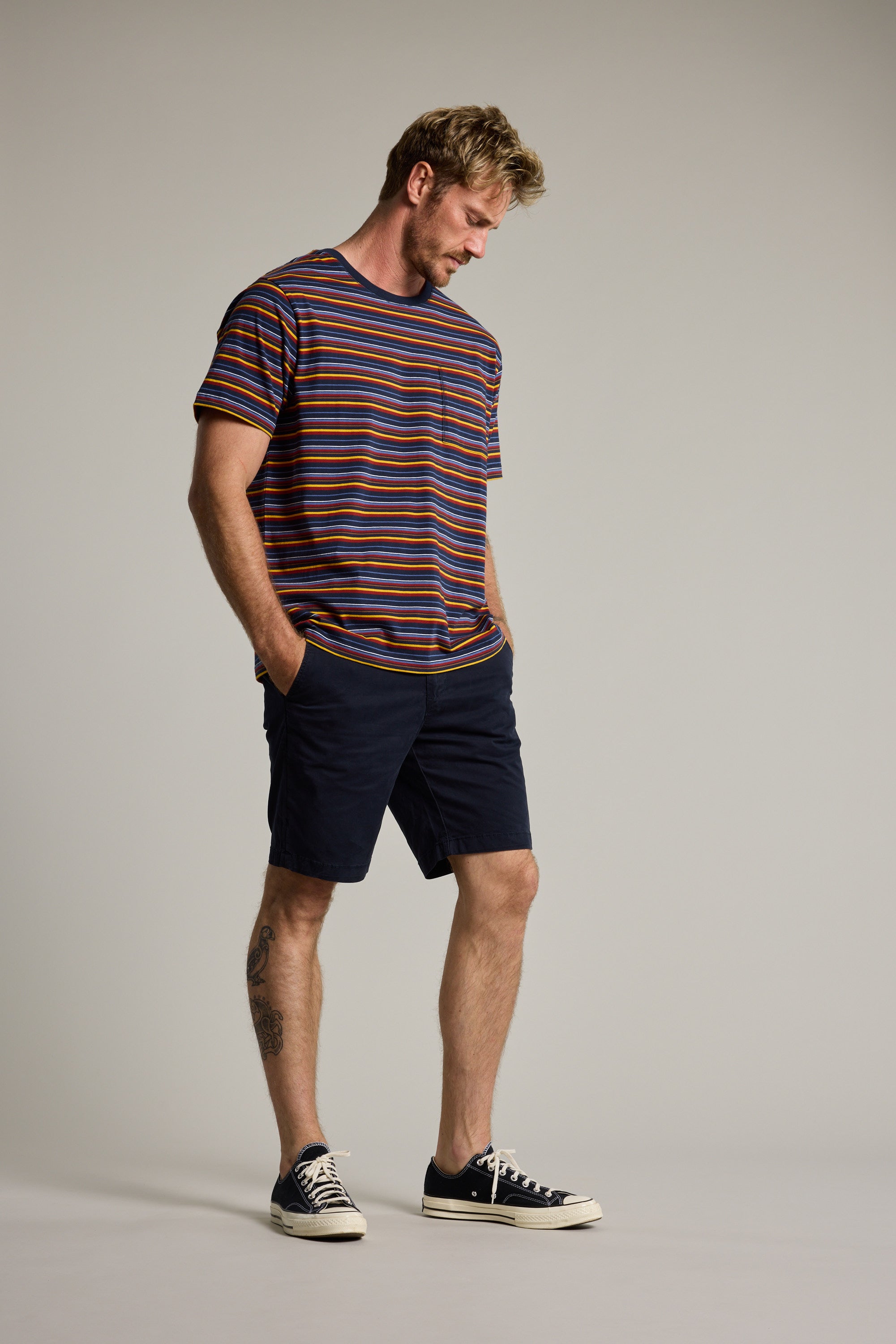 A man stands against a plain background in the Barkers Oxton Stripe Tee, crafted from organic cotton. He pairs it with navy shorts and black sneakers, standing relaxed with hands in pockets and a tattoo visible on his left calf.