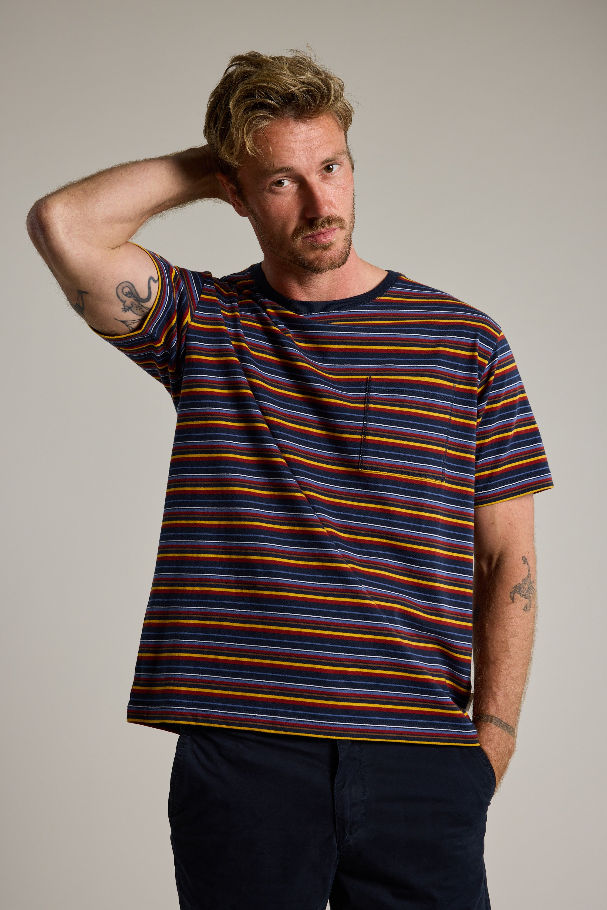 A man with blonde hair and tattoos wears the Barkers Oxton Stripe Tee, a men's regular fit tee with multicolored stripes and dark pants. He poses against a plain background, one hand behind his head, looking at the camera.