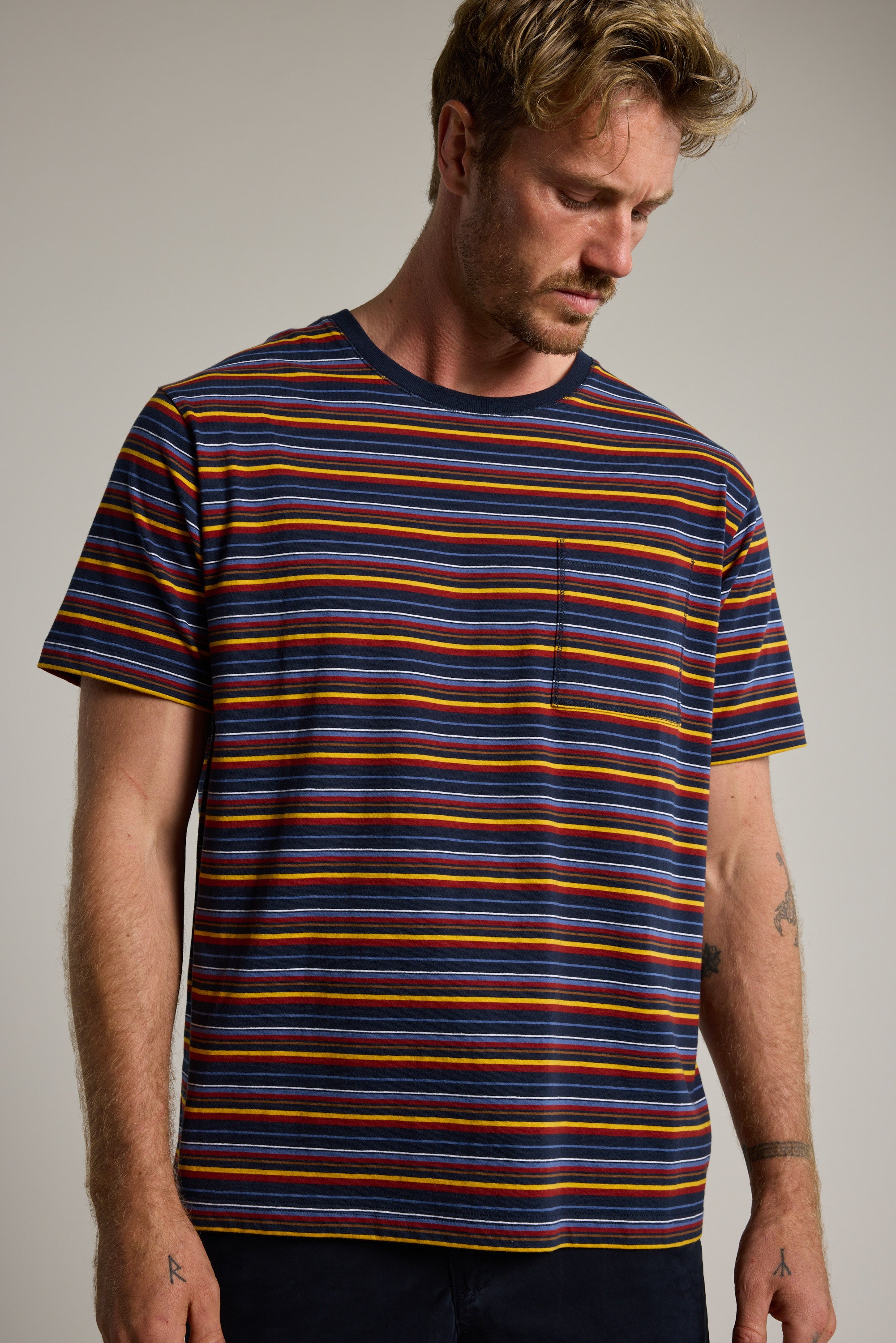 A man with light brown hair and a beard looks down while wearing the Barkers Oxton Stripe Tee, a multicolored striped, short-sleeved organic cotton shirt with a chest pocket, set against a neutral background.
