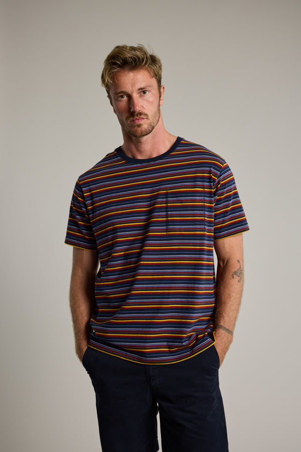 A blond man with a short beard stands against a plain background, wearing the Barkers Oxton Stripe Tee—an organic cotton men's regular fit tee with navy blue, orange, and brown stripes—dark pants, and showing a tattoo on his left forearm.