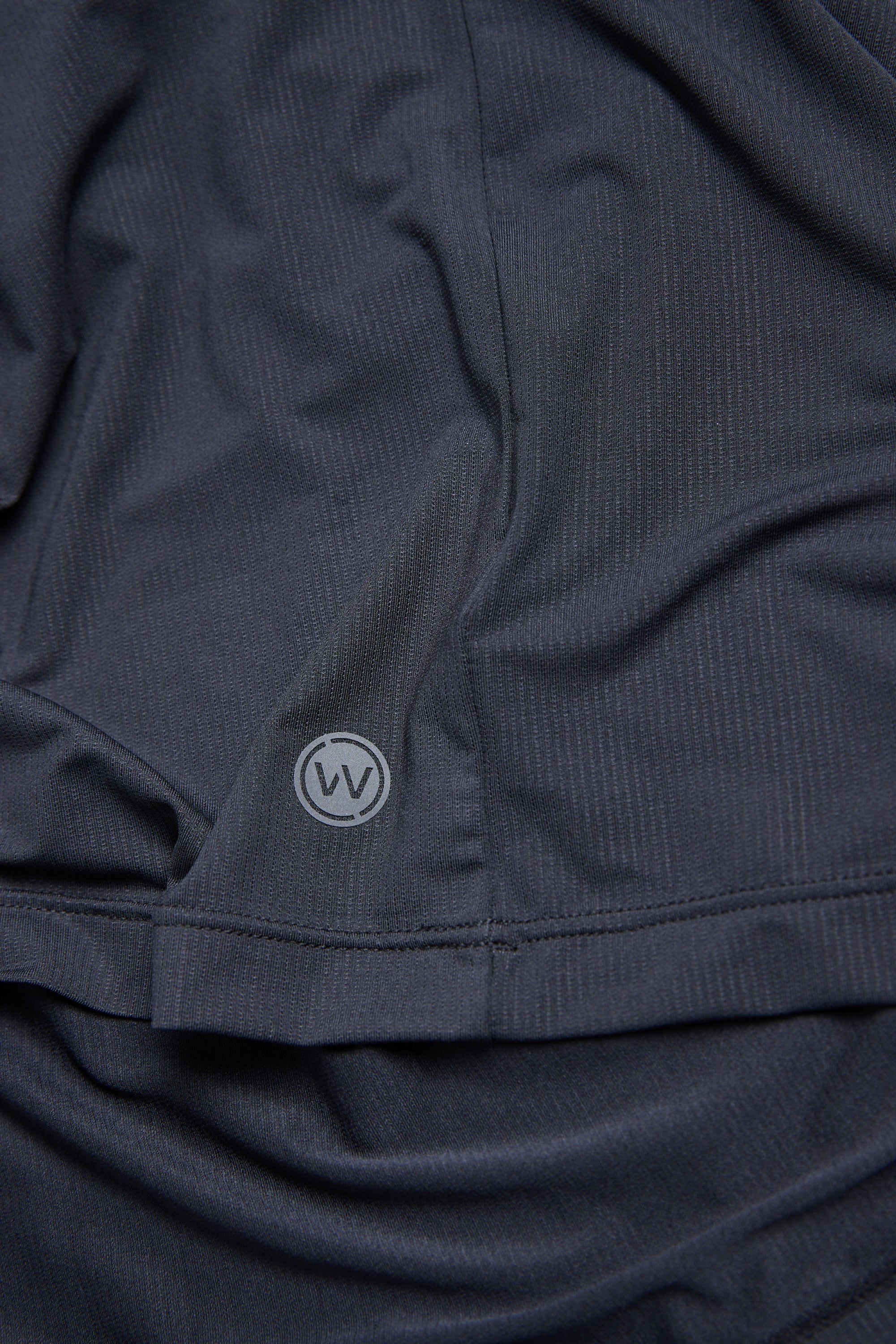 A close-up of the WearEver Pulse Long Sleeve Tee by WearEver in dark gray, featuring a textured fabric, subtle folds, a small circular WV logo near the hem, and UPF protection for sun safety.