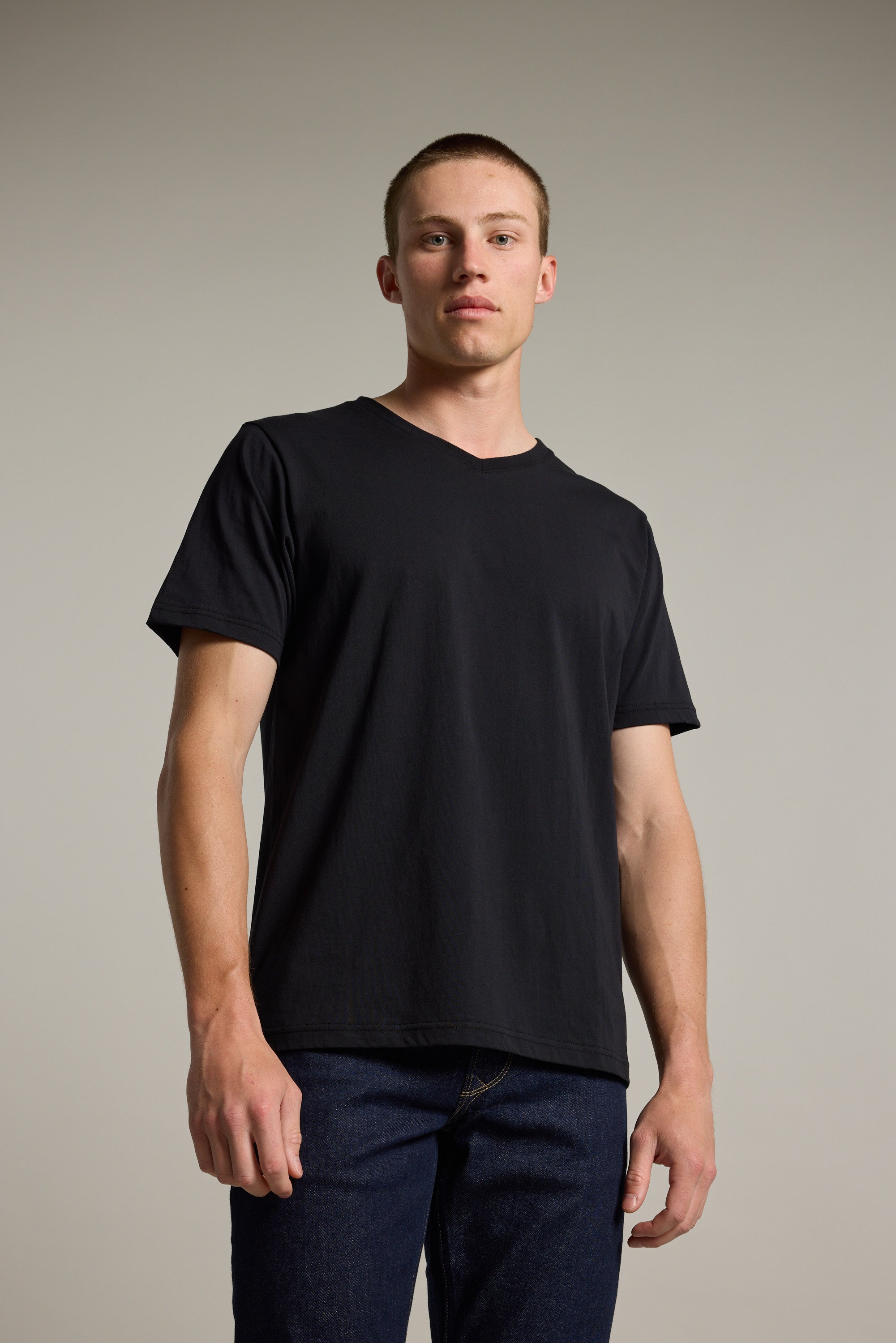 A young man stands against a plain background wearing the Barkers Organic Cotton V Neck Tee and dark jeans, confidently facing the camera with relaxed arms—a comfortable, everyday essential for effortless style.