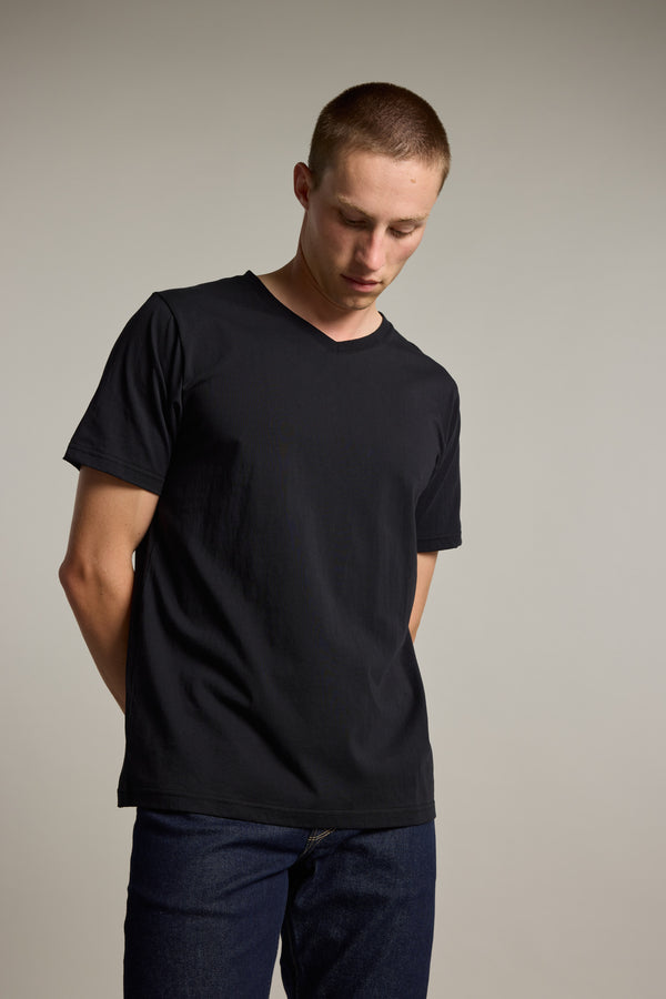 A young man with short hair stands against a neutral background, hands behind his back and looking down, wearing the Barkers Organic Cotton V Neck Tee paired with dark jeans.