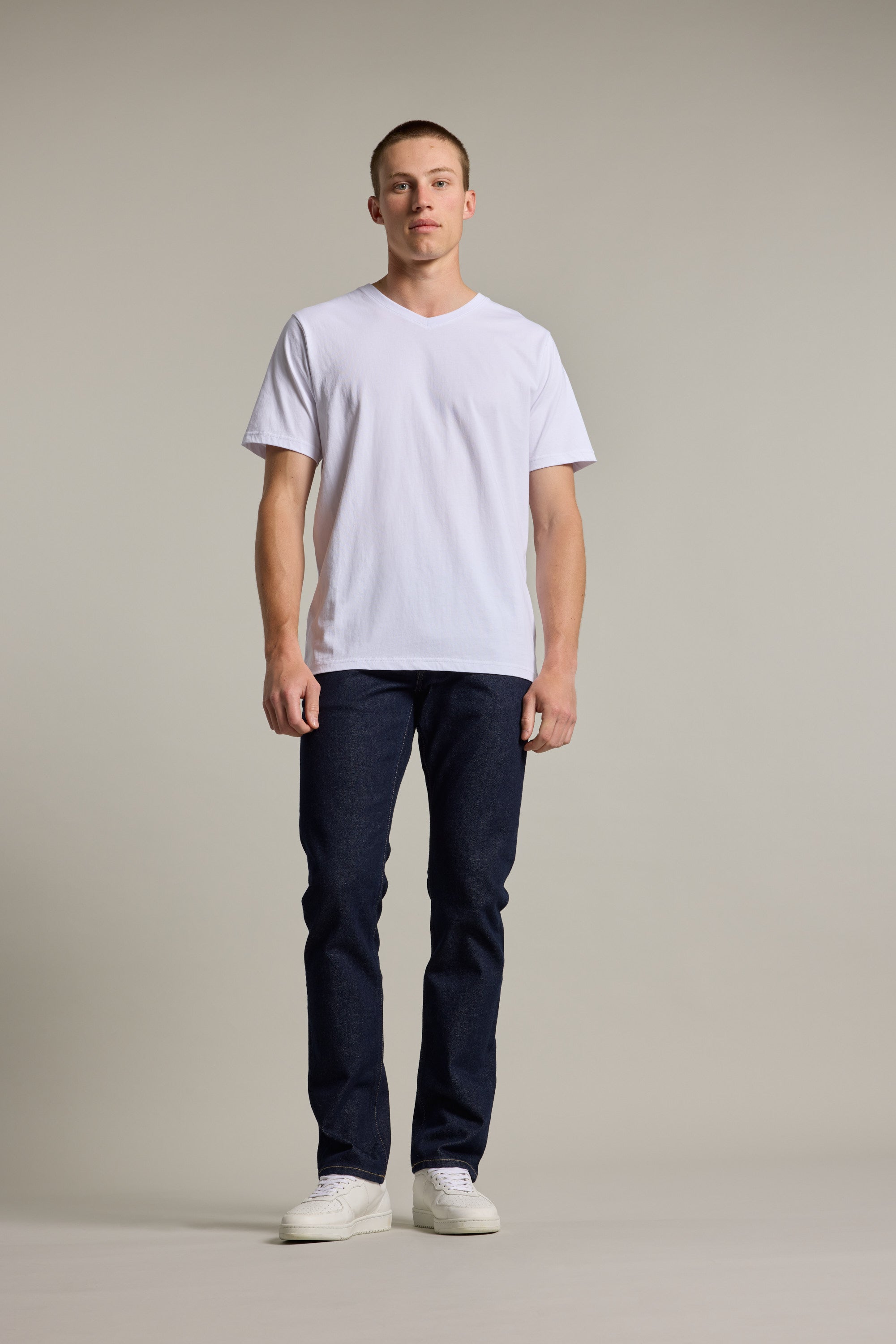 A man with short hair and a neutral expression faces forward against a plain light background, wearing the Barkers Organic Cotton V Neck Tee, paired with dark blue jeans and white sneakers.