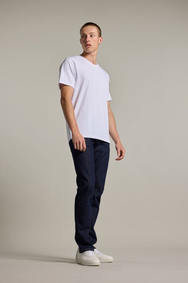A young man stands against a plain background in the Barkers Organic Cotton V Neck Tee, paired with dark blue jeans and white sneakers. He gazes slightly to the side with a neutral expression.