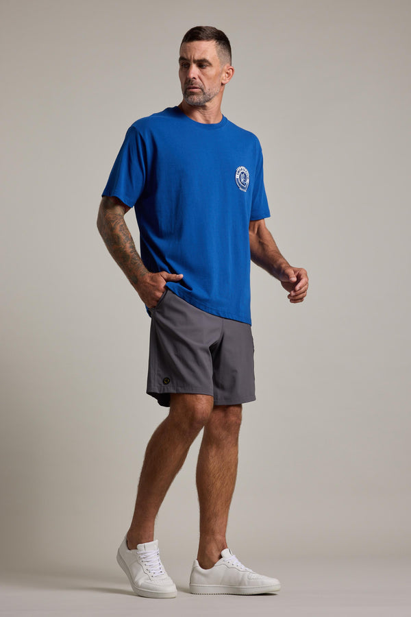 A man stands against a plain background, wearing a blue Swordfish Mojo Logo Tee by Swordfish Mojo, gray shorts, and white sneakers. He has short hair, a beard, visible arm tattoos, and looks slightly to the side with one hand in his pocket.