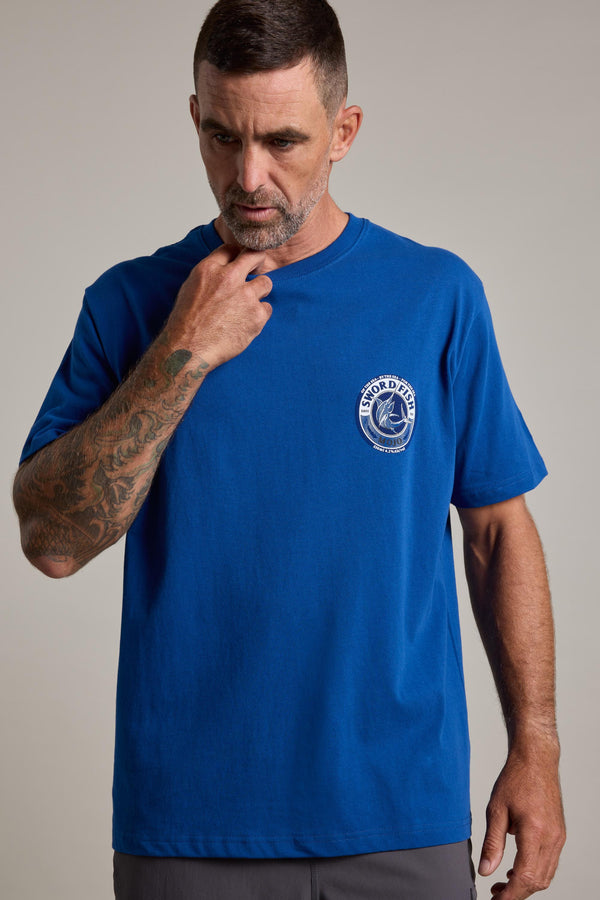 A man with short hair and tattoos on his left arm wears a blue Swordfish Mojo Logo Tee by Swordfish Mojo, featuring a white circular logo on the chest. He looks down and touches his neck with his right hand.