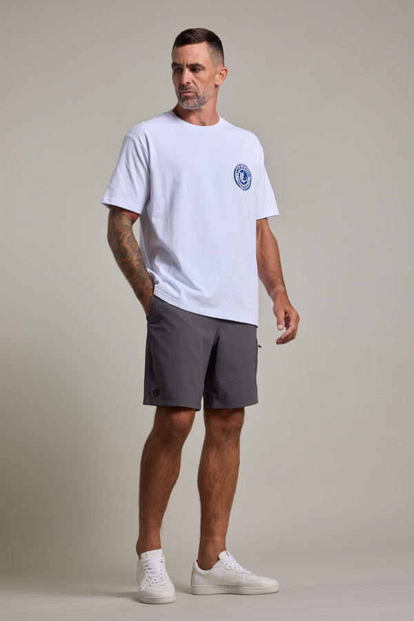A man with short hair and a trimmed beard stands against a plain background wearing the Swordfish Mojo Logo Tee by Swordfish Mojo—an exclusive white organic cotton shirt with a blue emblem—paired with gray shorts and white sneakers, one hand in his pocket.