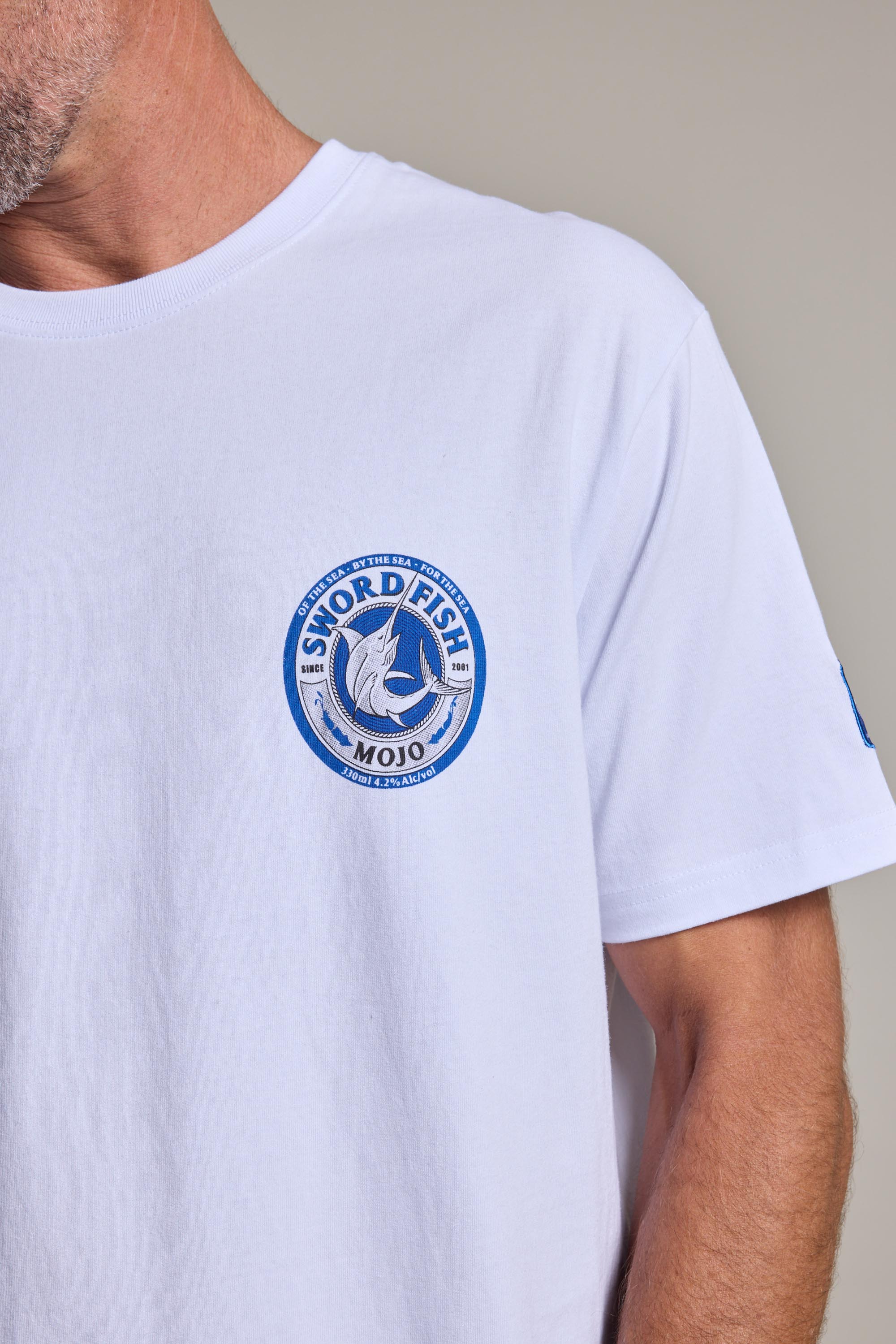 A person wears the Swordfish Mojo Logo Tee, an organic cotton shirt from Swordfish Mojo, featuring a blue circular chest logo with a swordfish and “Sword Fish Mojo” text; their head is partially out of frame.