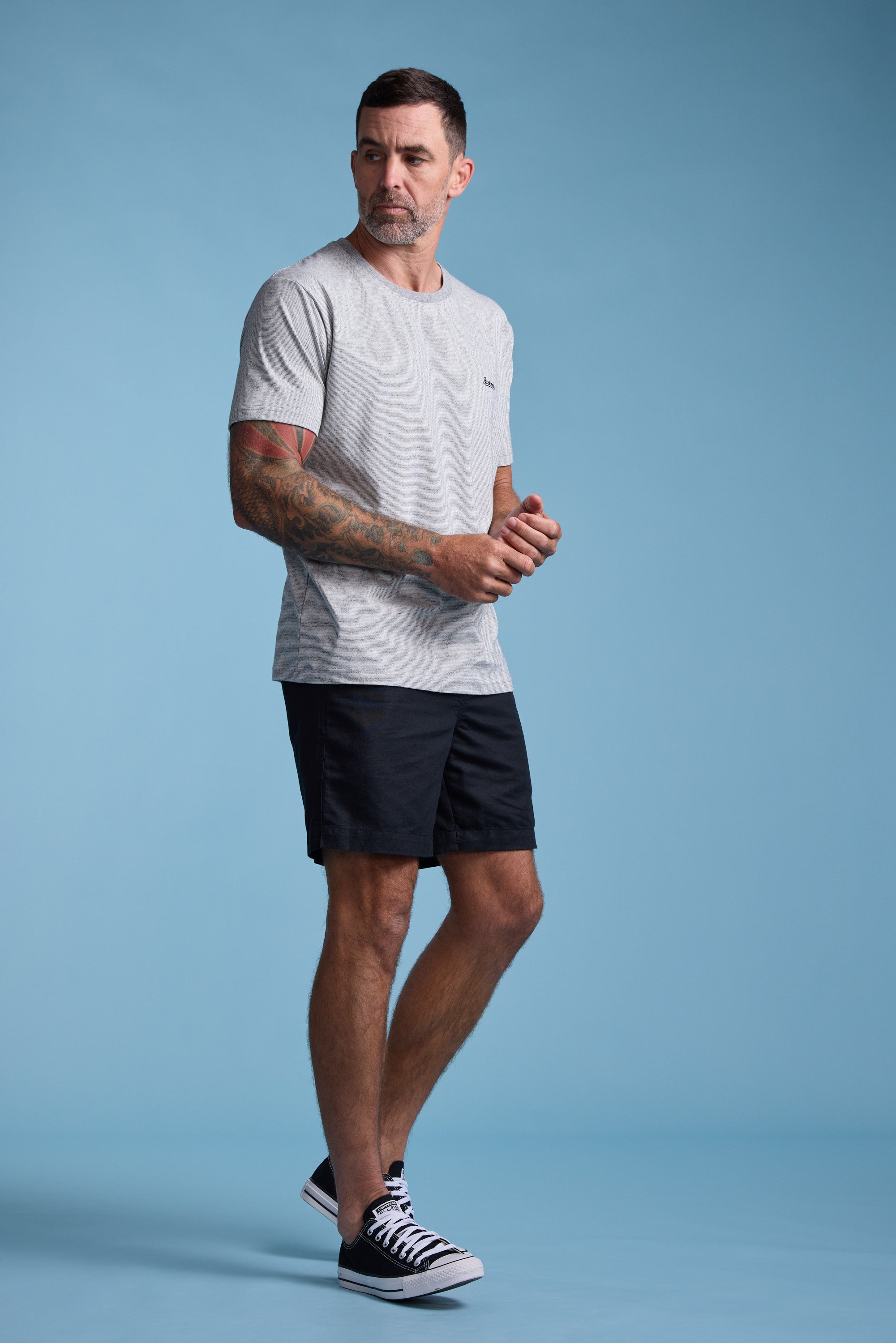 A man with short dark hair and tattooed arms stands against a light blue background, wearing the Bayside Emb Tee by Barkers in light gray, black shorts, and black sneakers with white soles, looking slightly to his left.