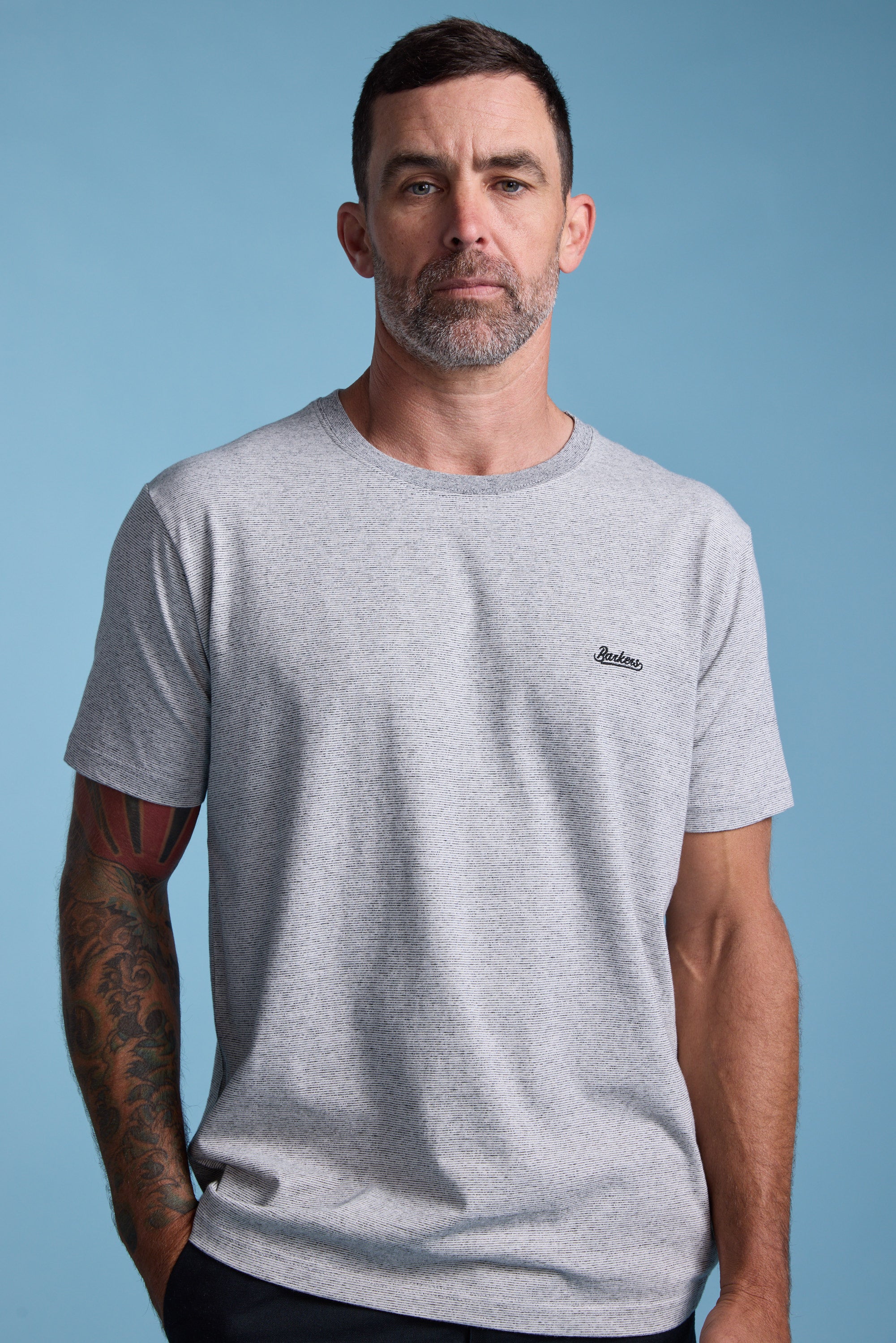 A man with short dark hair and a beard wears the Barkers Bayside Emb Tee, a light grey organic cotton T-shirt with a small embroidered logo on the chest, standing against a plain blue background. Tattoo visible on his left forearm.