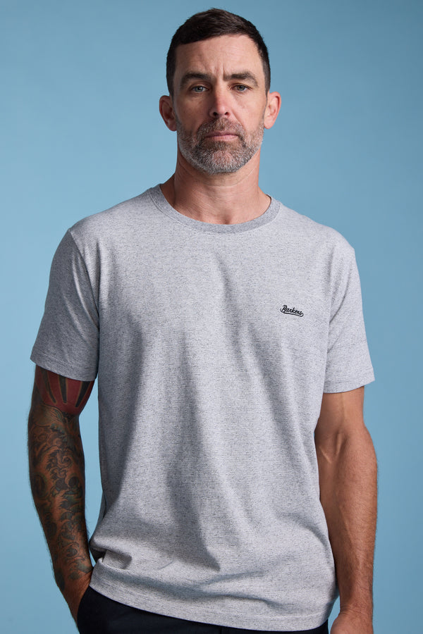 A man with short dark hair and a beard wears the Barkers Bayside Emb Tee, a light grey organic cotton T-shirt with a small embroidered logo on the chest, standing against a plain blue background. Tattoo visible on his left forearm.