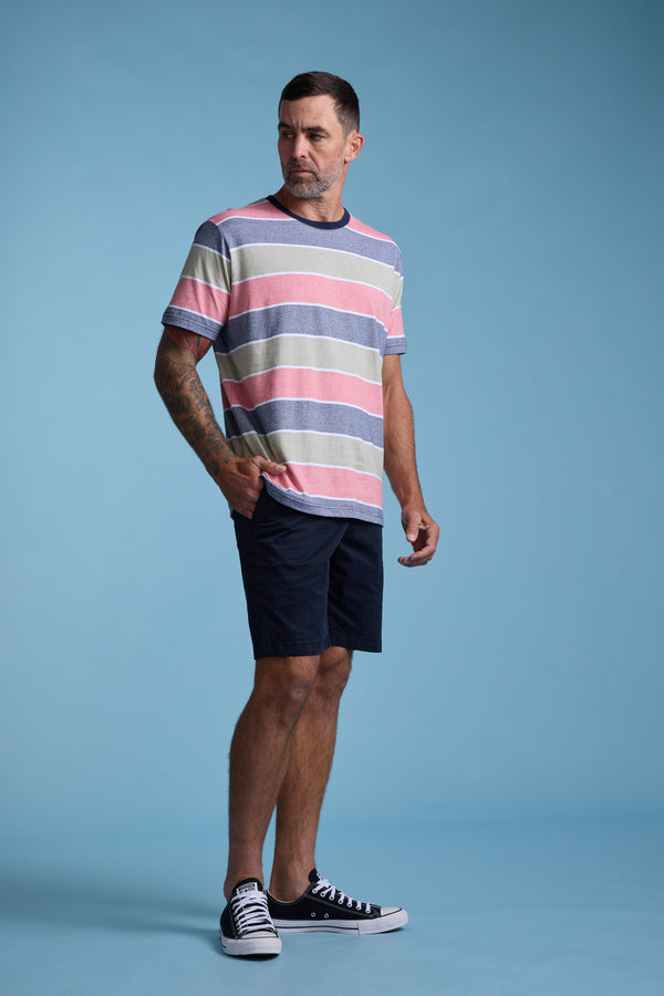 A man with short hair and visible left-arm tattoos stands against a blue background wearing the Barkers Asbrooke Stripe Tee, dark shorts, and black sneakers. He looks to his right with a neutral expression.