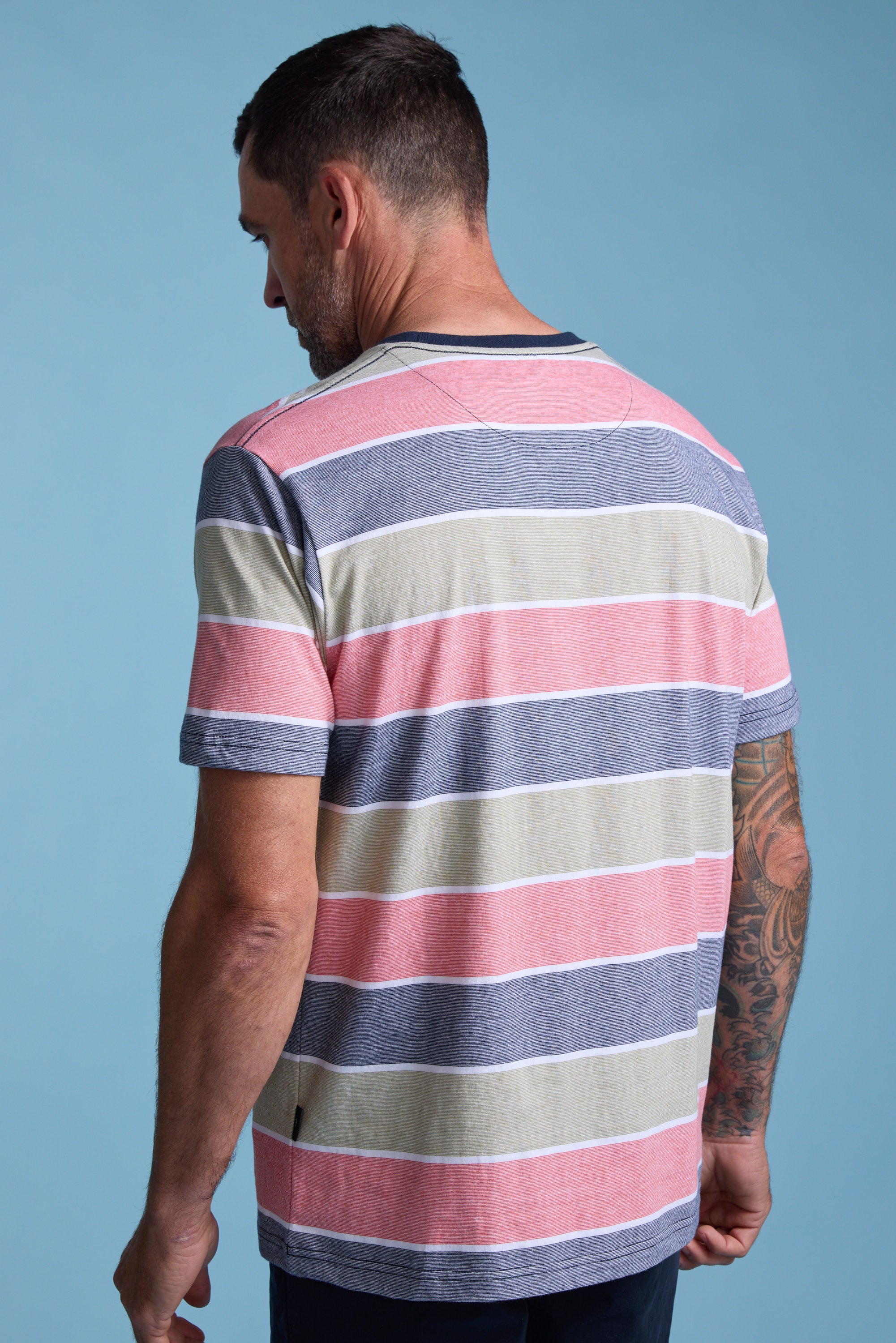 A man with short dark hair and a tattooed right arm stands with his back to the camera, wearing the Barkers Asbrooke Stripe Tee—an organic cotton t-shirt in pink, beige, and gray stripes—set against a blue background.