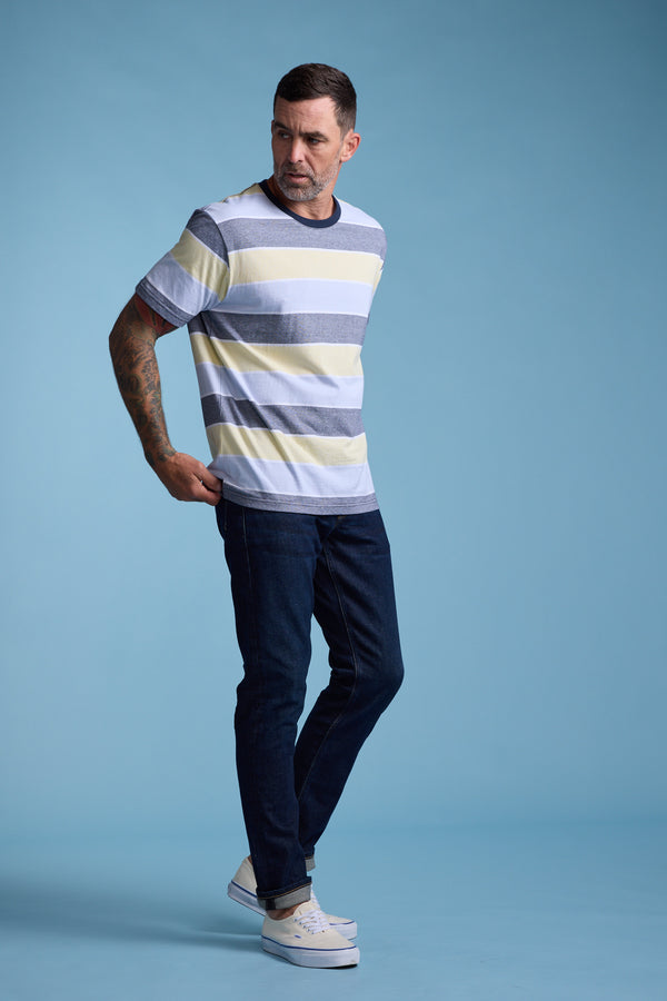 A man stands against a light blue background, wearing the Barkers Asbrooke Stripe Tee in gray, white, and yellow stripes, paired with dark blue jeans and white sneakers. He looks to his left with one hand behind his back.