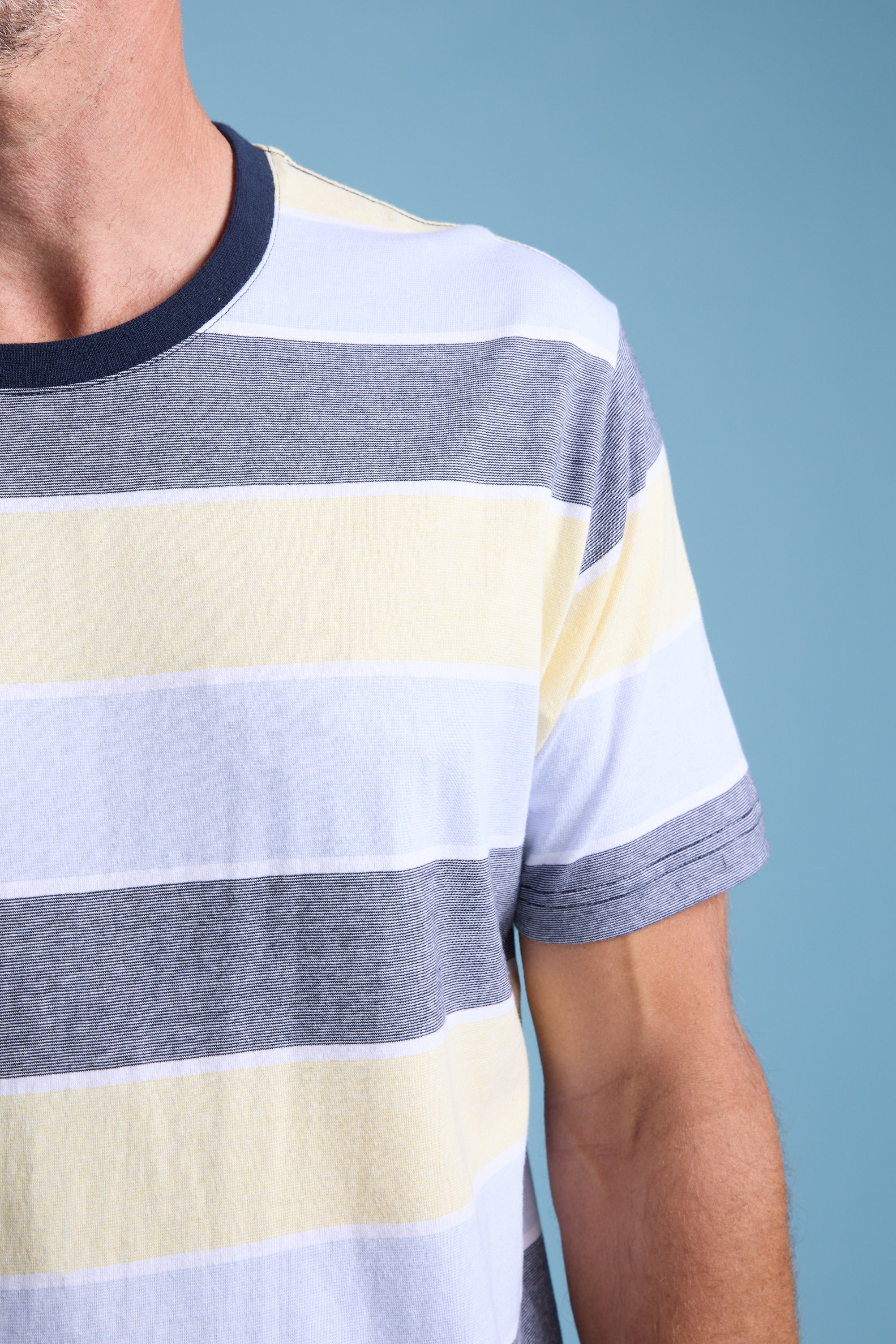 A close-up of a person wearing the Barkers Asbrooke Stripe Tee—a regular fit, short-sleeve crew neck organic cotton top with white, yellow, and gray horizontal summer stripes—shows only the upper torso and left arm against a light blue background.
