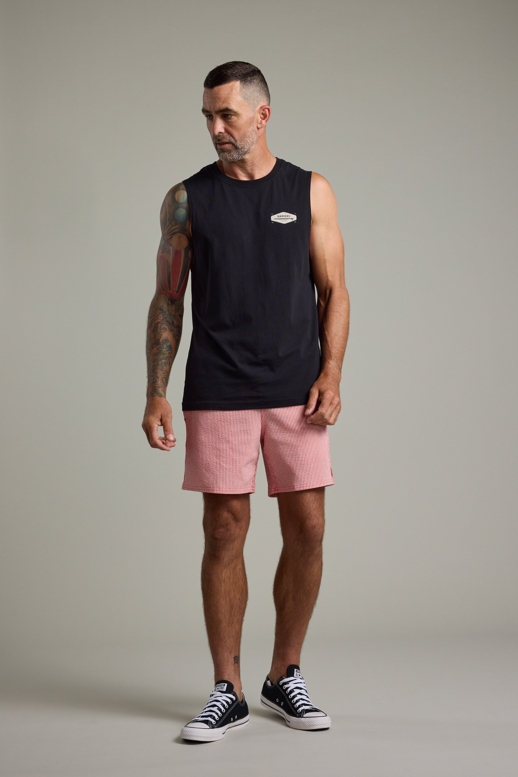 A man with short dark hair and a beard wears a sleeveless black shirt, Barkers Dehaven Stripe Volley shorts, and black sneakers. He has a colorful arm tattoo and stands against a plain light background, looking slightly to his left.
