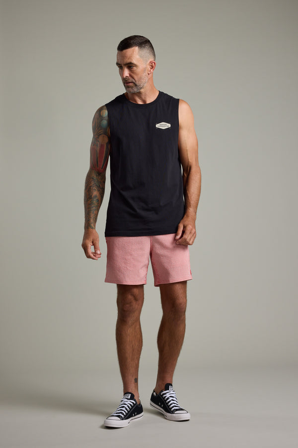 A man with short dark hair and a beard wears a sleeveless black shirt, Barkers Dehaven Stripe Volley shorts, and black sneakers. He has a colorful arm tattoo and stands against a plain light background, looking slightly to his left.