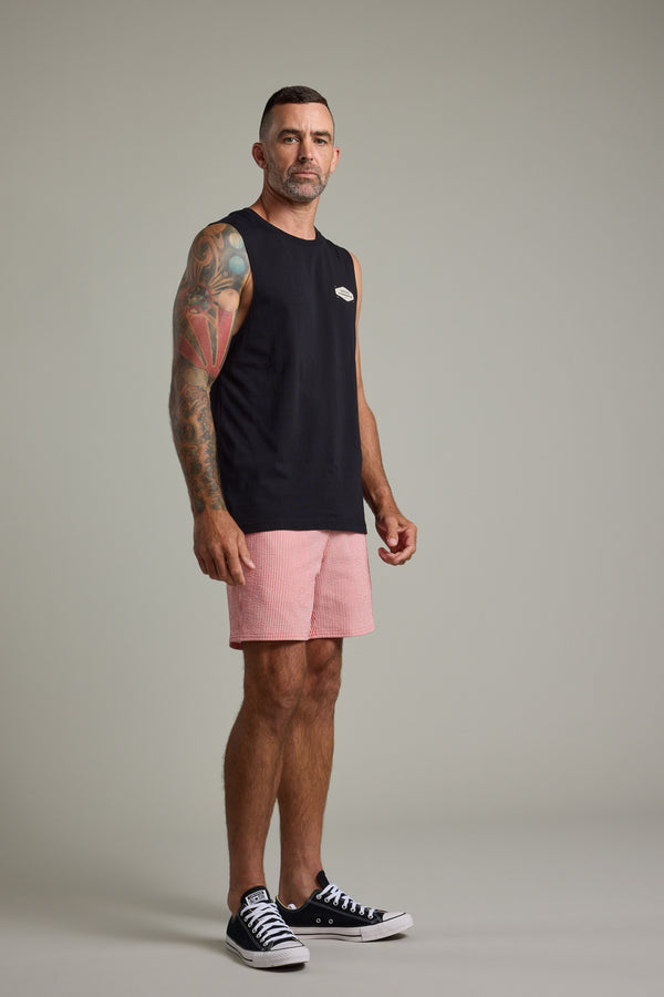 A man with short hair and arm tattoos stands against a plain backdrop, wearing the Barkers Supply Co Print Singlet and pink striped Barkers longboard shorts with black sneakers—an ideal summer look.