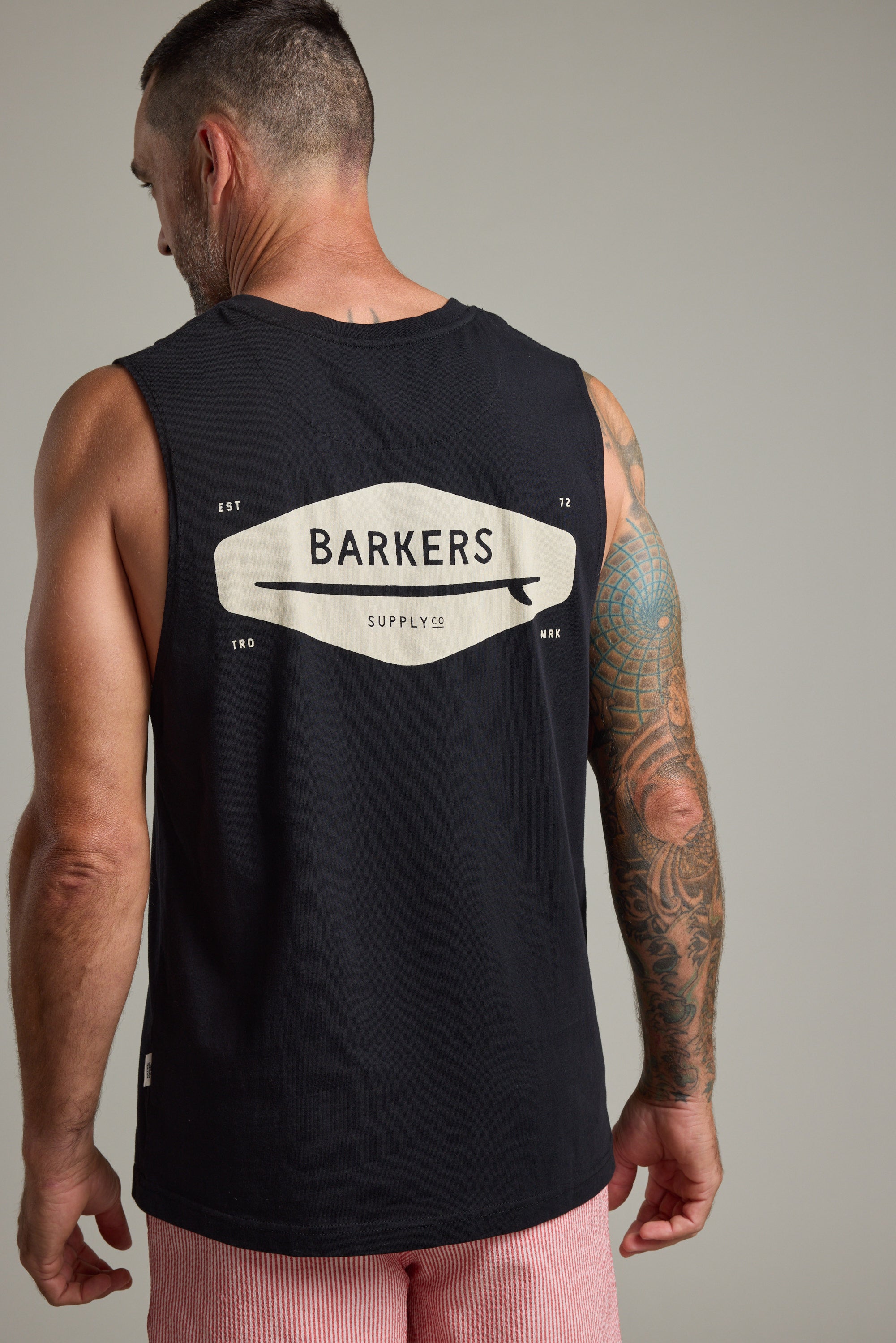 A man with short hair and a tattooed right arm wears the black Barkers Supply Co Print Singlet with logo on the back, facing away from the camera in pink and white striped shorts—a true summer essential by Barkers.