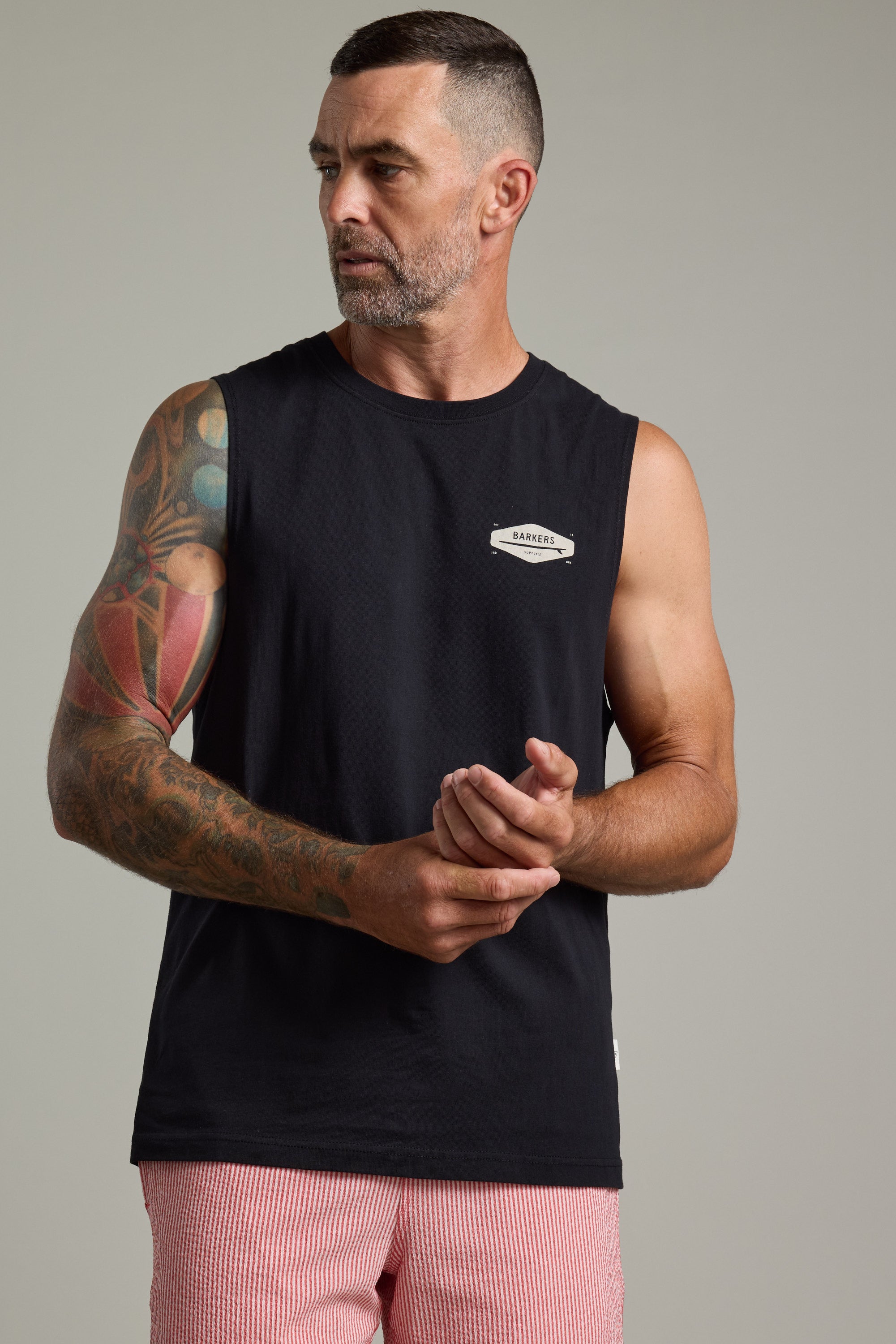A man with short hair and a beard wears the Barkers Supply Co Print Singlet and red-and-white striped shorts. Sporting a colorful tattoo sleeve, he looks left while adjusting his right wrist, ready for a summer-essential Barkers longboard ride.
